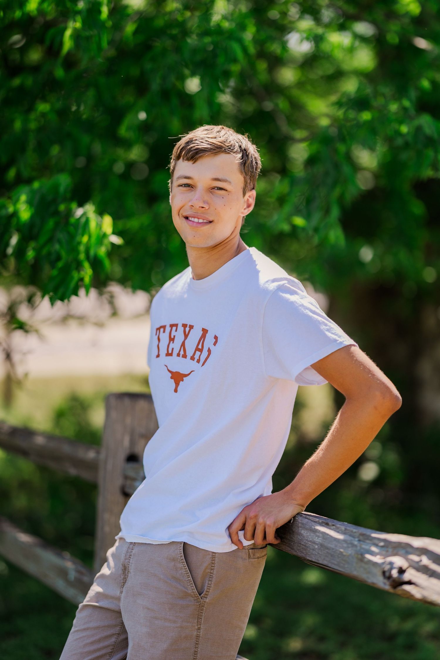 Smiling teen boy in white Texas Longhorns t-shirt leans on wooden fence surrounded by green trees.