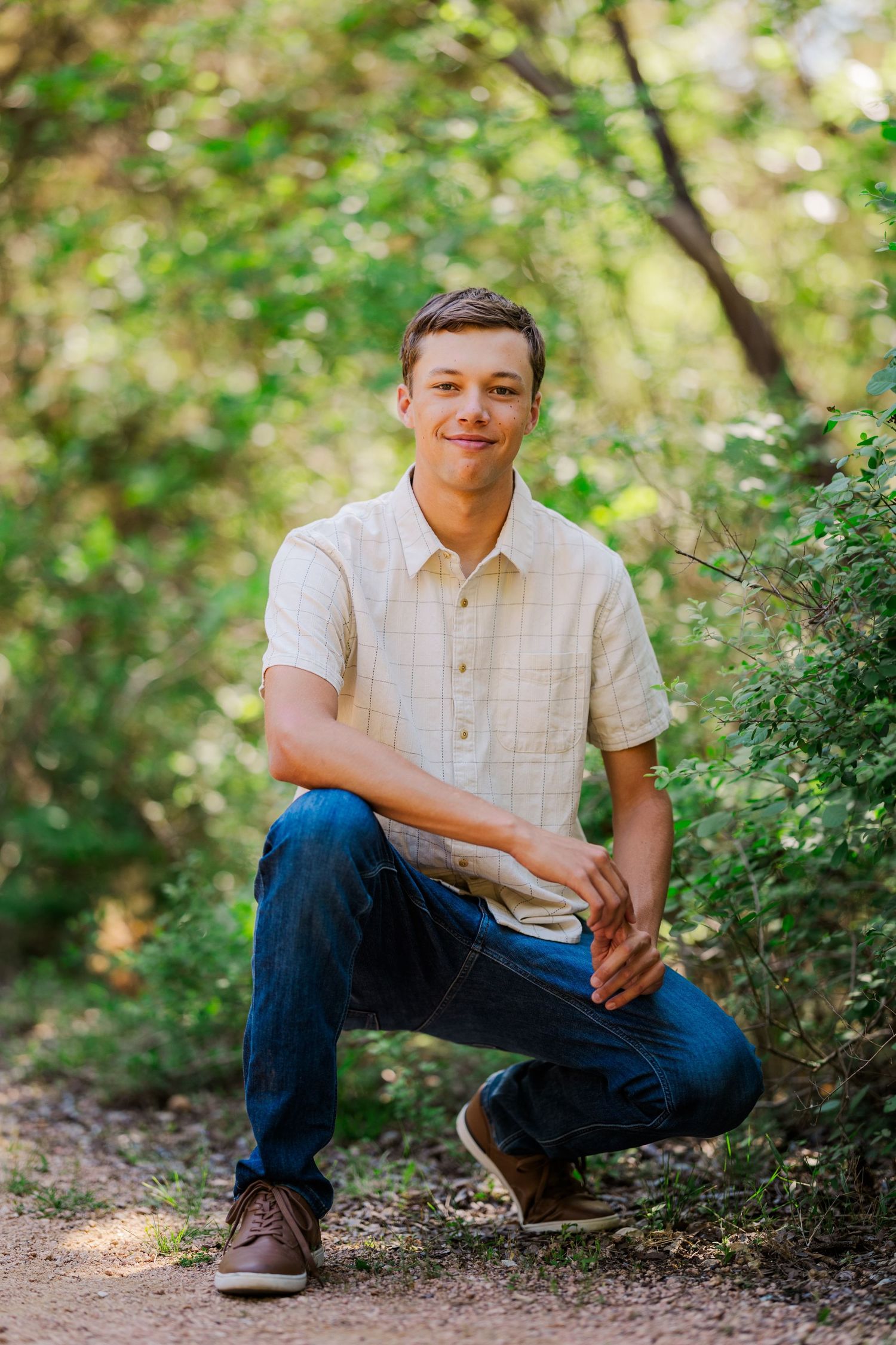 Young man in beige shirt and jeans crouching on forest path surrounded by green trees.