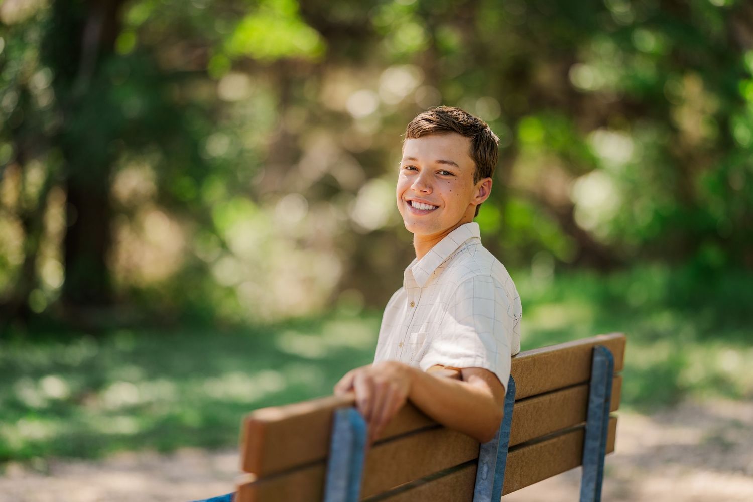 Smiling teenage boy sitting on a wooden park bench surrounded by green trees.