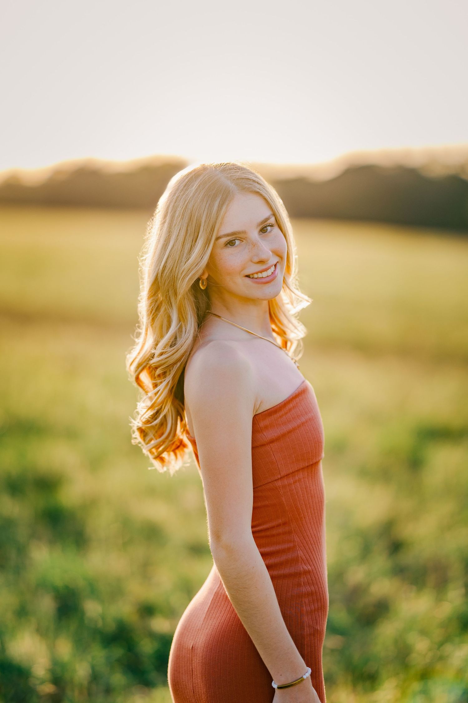 Blonde teenage girl in rust orange dress posing in golden hour sunlit field for senior portrait.
