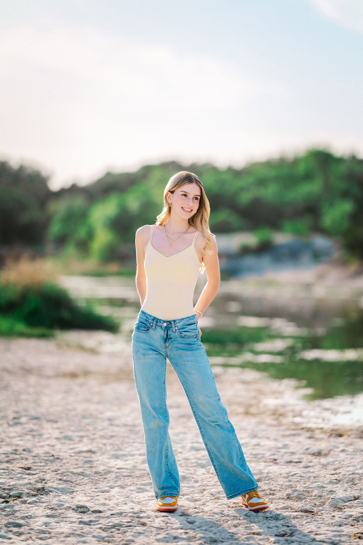 Smiling blonde young woman in cream tank and wide-leg jeans stands on a sandy riverbank at dusk.