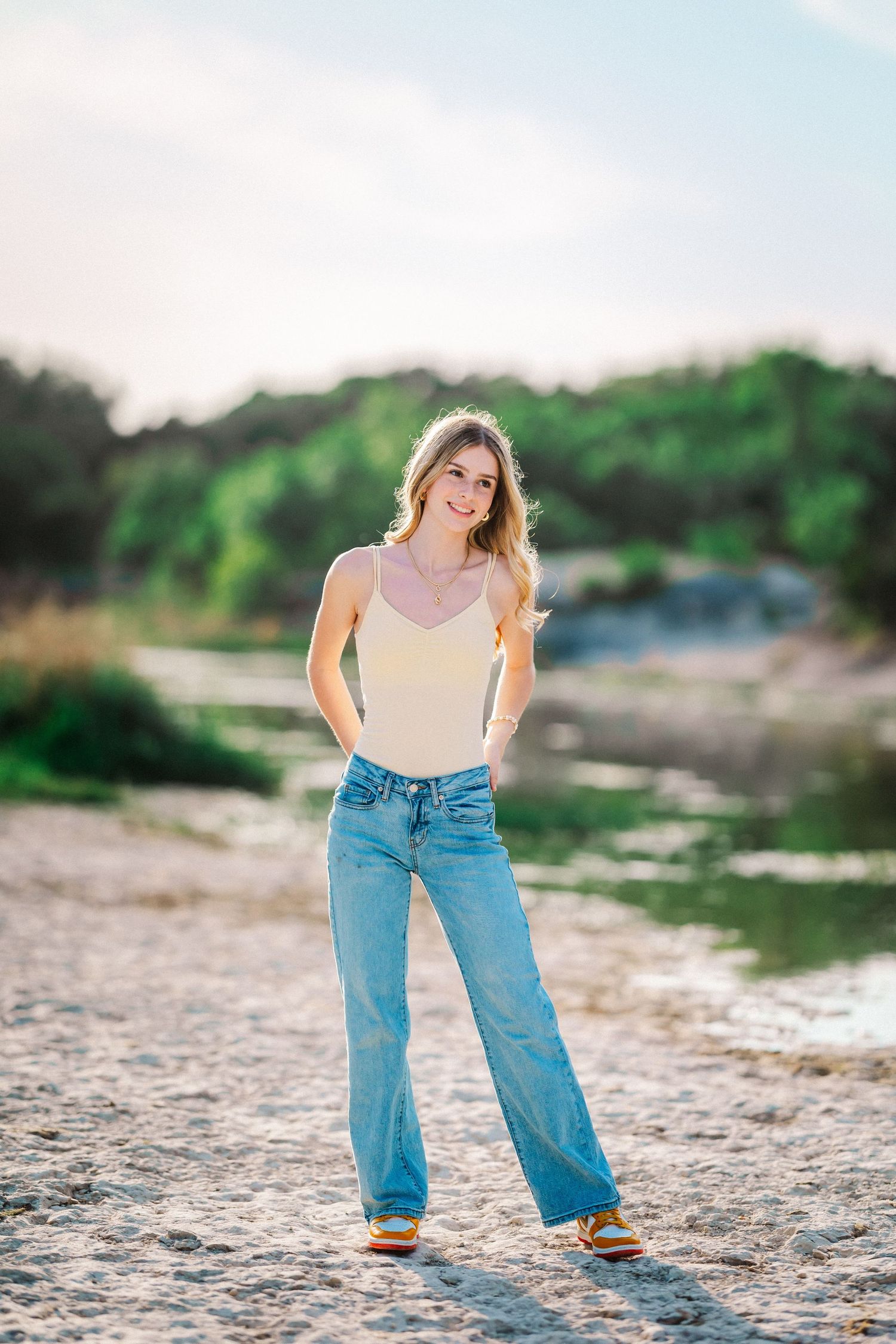 Smiling blonde teen in cream tank top and wide-leg jeans poses on a sandy riverbank at golden hour.
