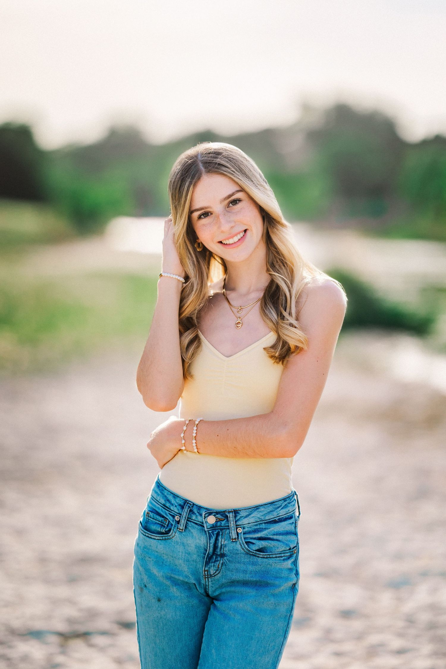 Smiling blonde teen in yellow tank top and jeans poses outdoors during golden hour on a gravel path.
