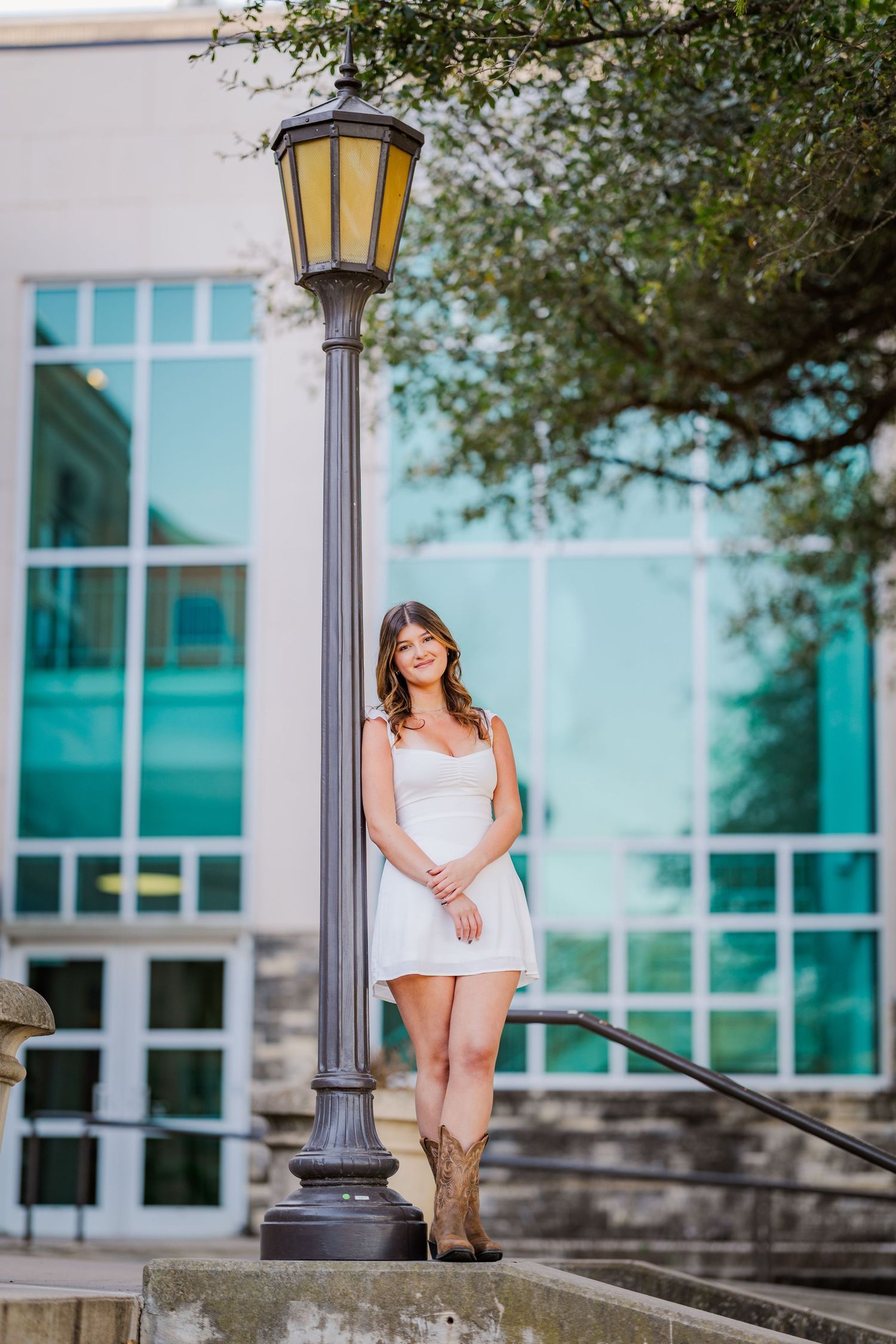 Young woman in white dress and cowboy boots posing by lamppost outside modern building.