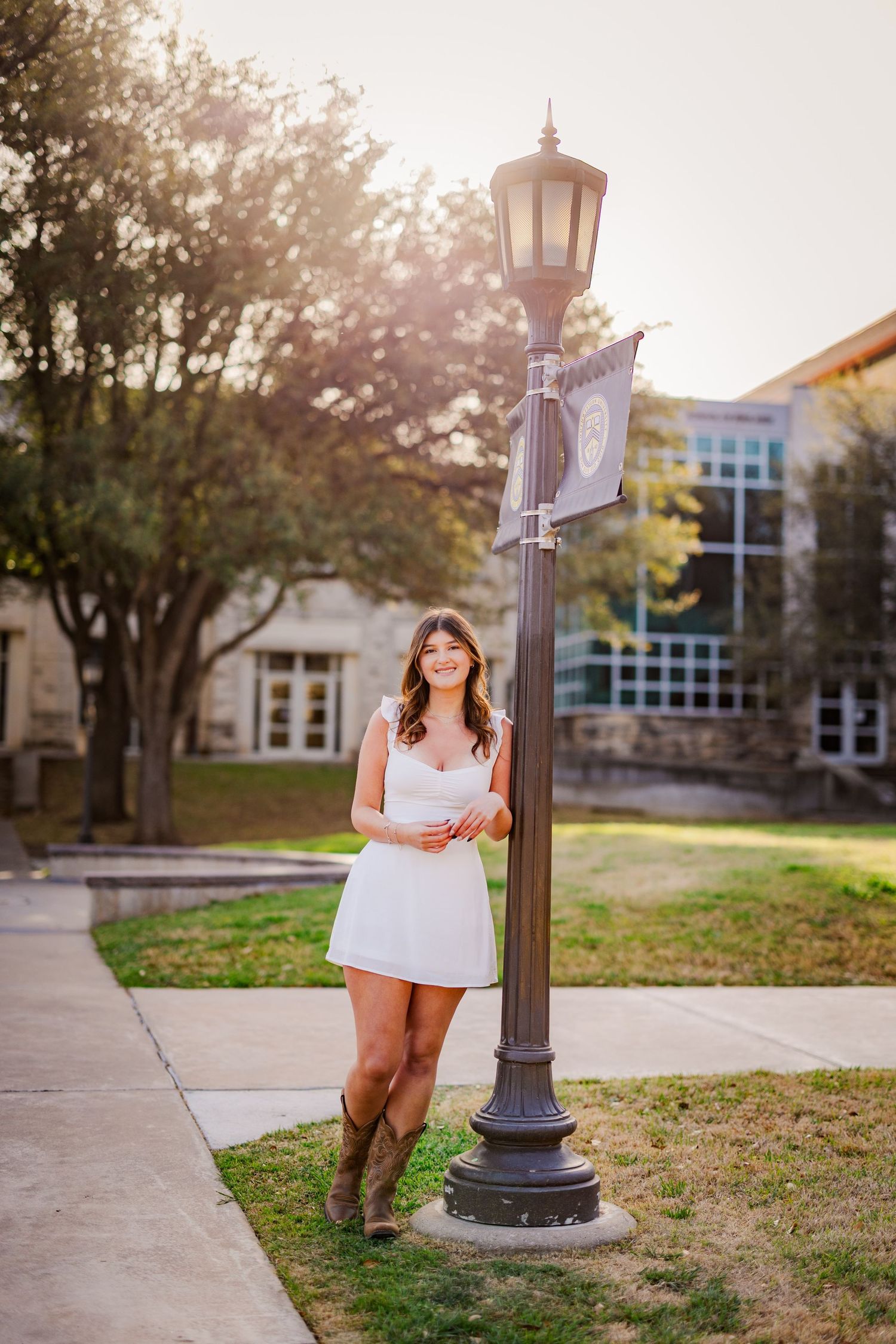 Young woman in white dress and cowboy boots leans against a lamp post on a sunny college campus.