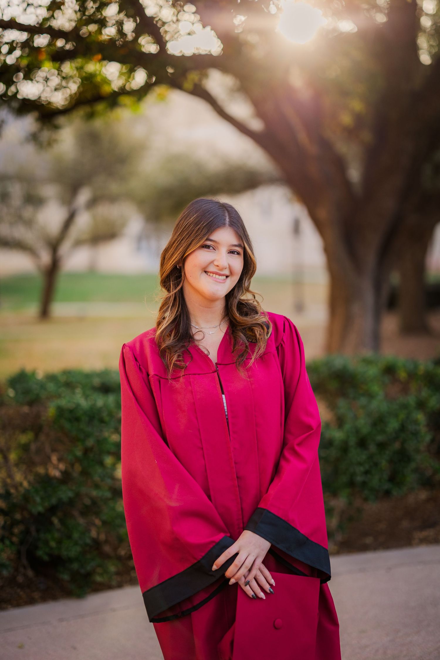 Smiling young woman in red graduation cap and gown posing outdoors at sunset.