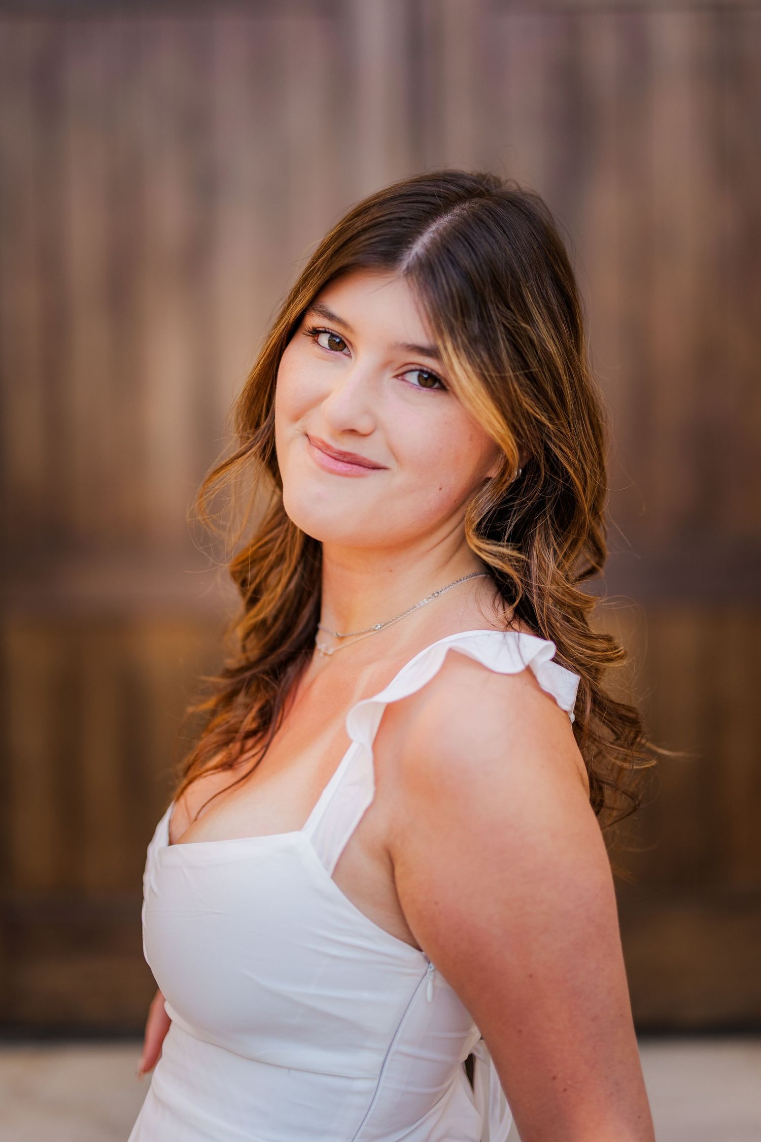Young woman with wavy brown hair smiles softly in a white dress against a wooden backdrop.