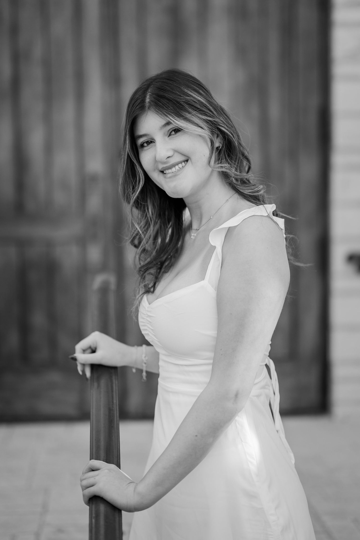 Young woman in white sundress smiles while holding a wooden railing in black and white portrait.