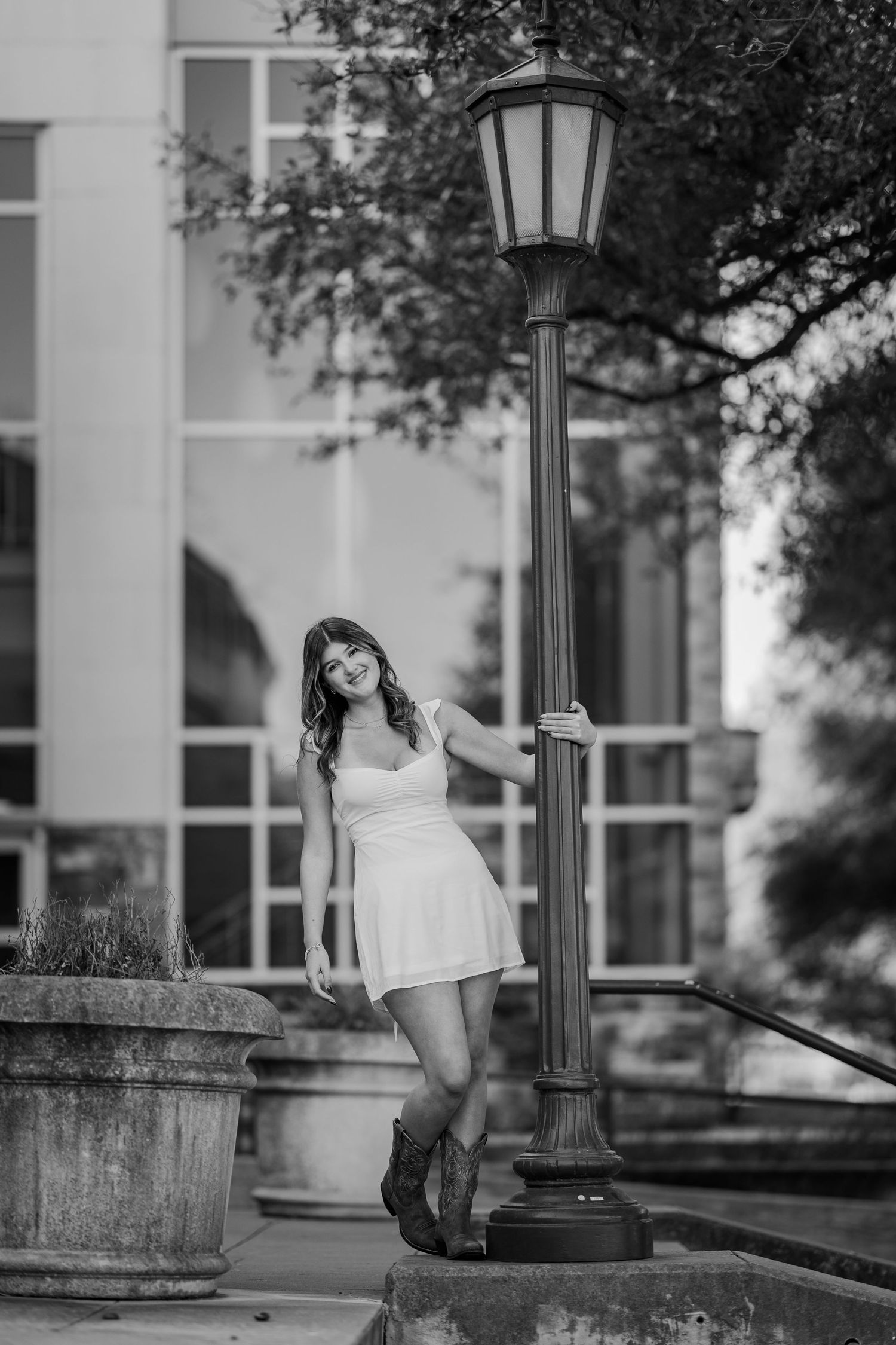 Black and white portrait of a teen girl in white dress leaning on a lamp post outside a building.