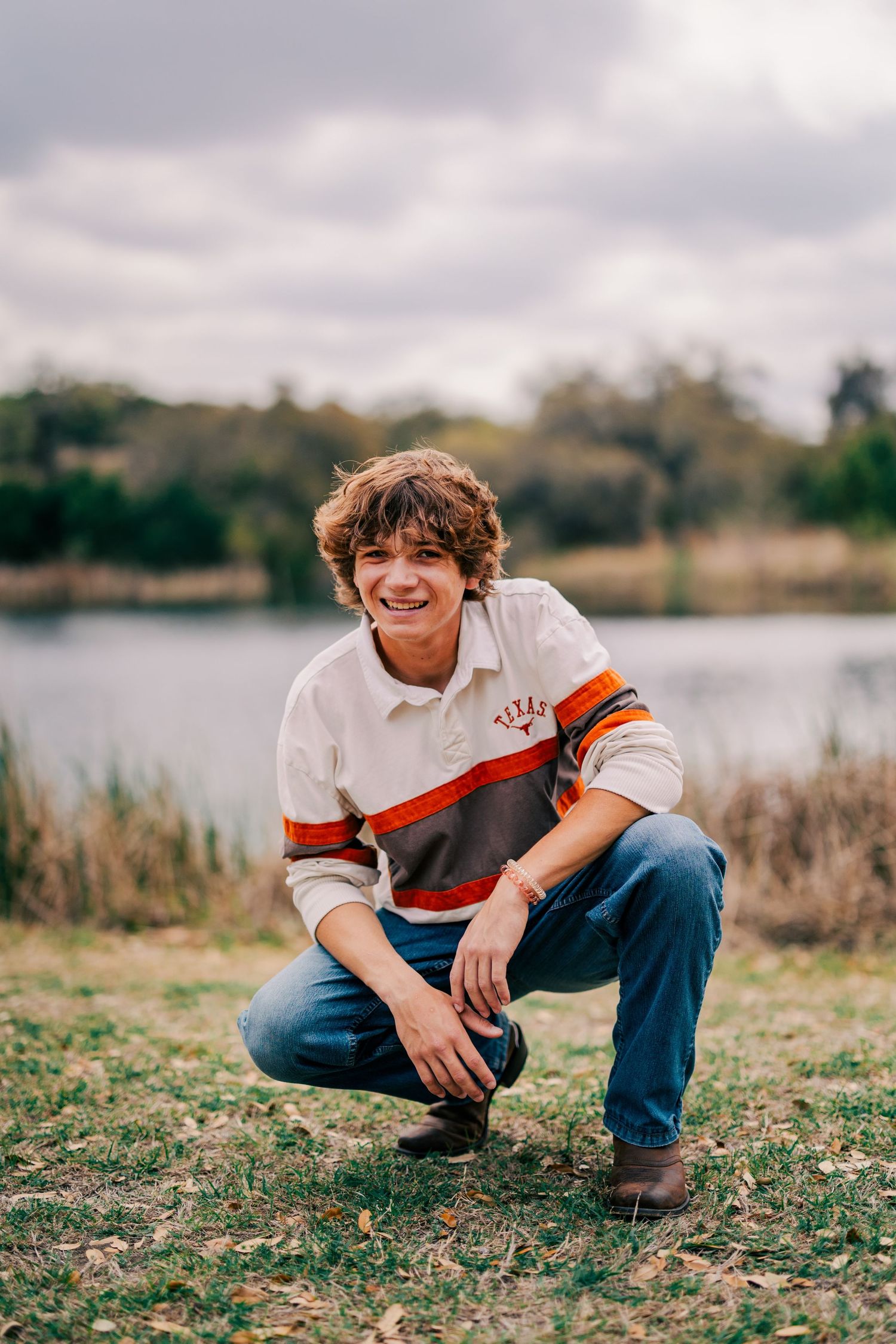 Smiling teen boy in Texas striped polo crouches on grass near a calm lake on cloudy day.