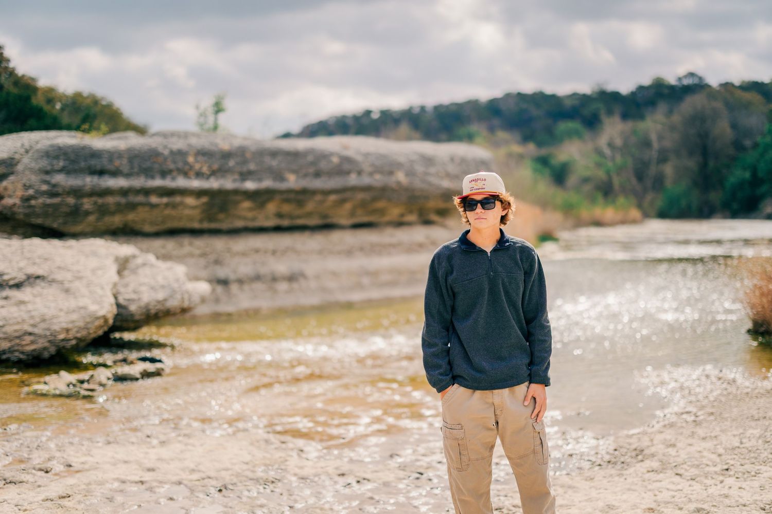 Young person in navy pullover and hat stands on rocky riverbed with cloudy sky and trees behind.