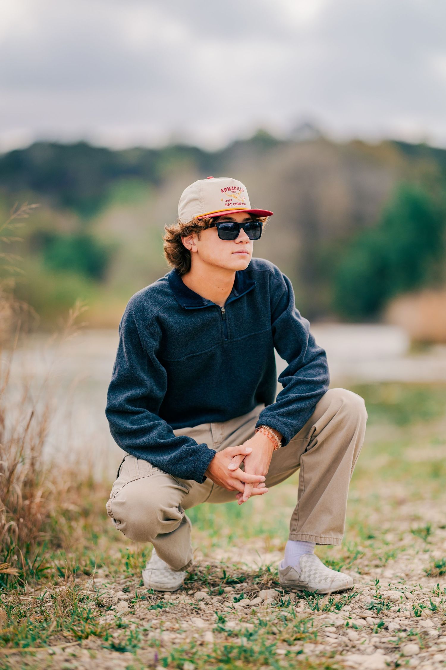 Teen boy in navy fleece and cap crouching outdoors near a lake in nature.