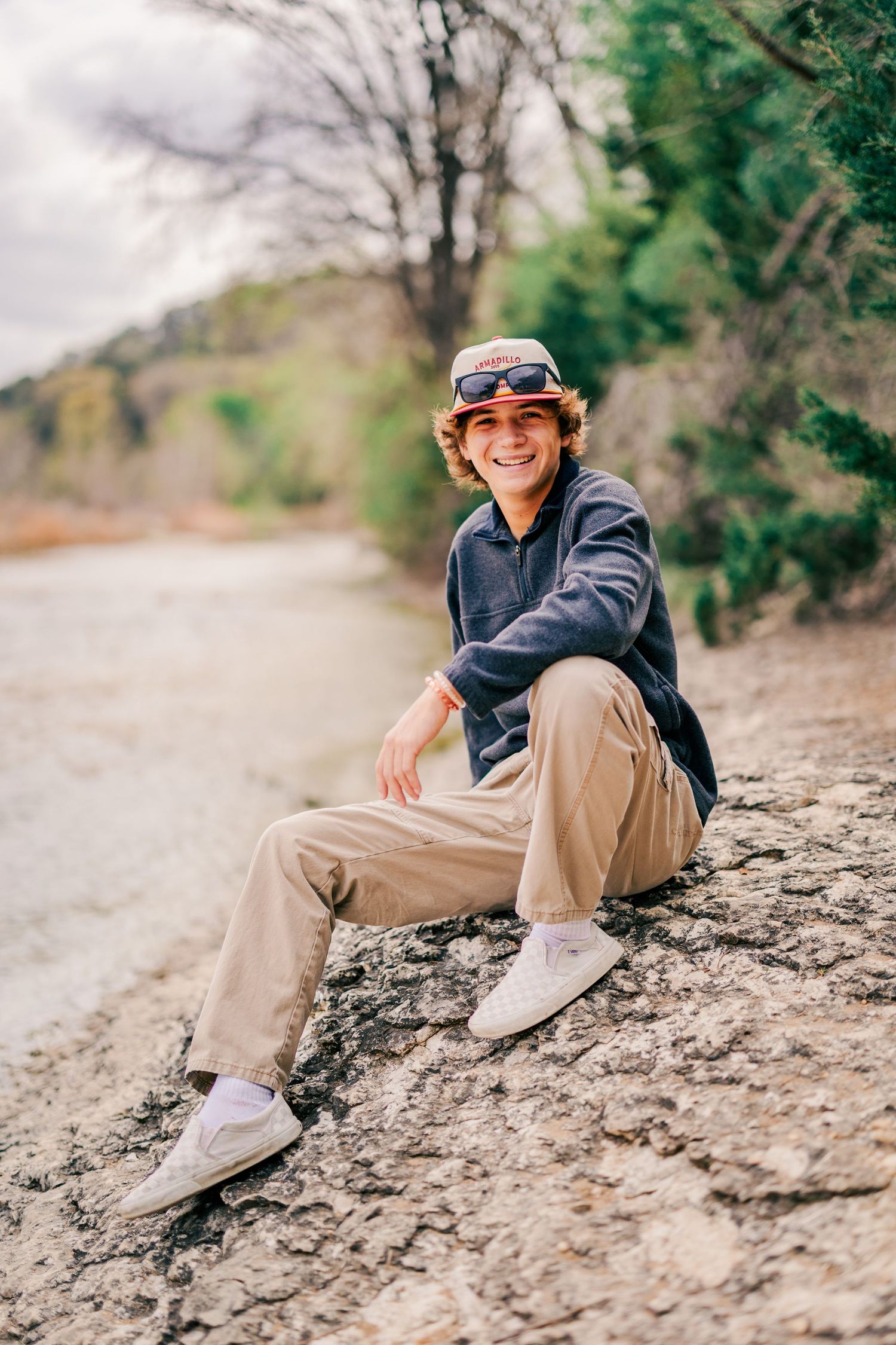 Smiling teen boy with sunglasses on cap sits on rocky trail surrounded by trees in nature.