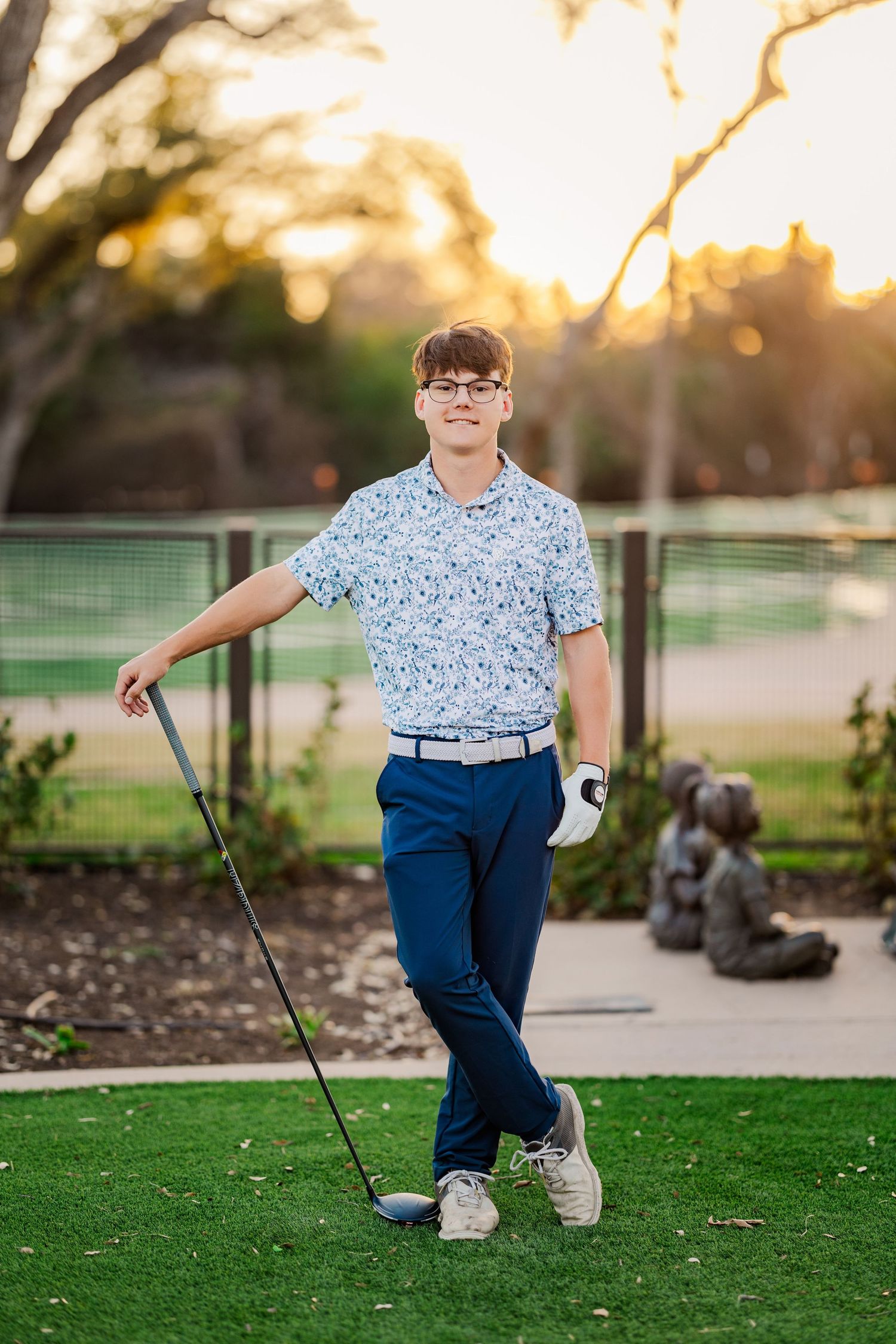 Young man in floral golf shirt leans on a golf club at a golf course during a warm sunset.