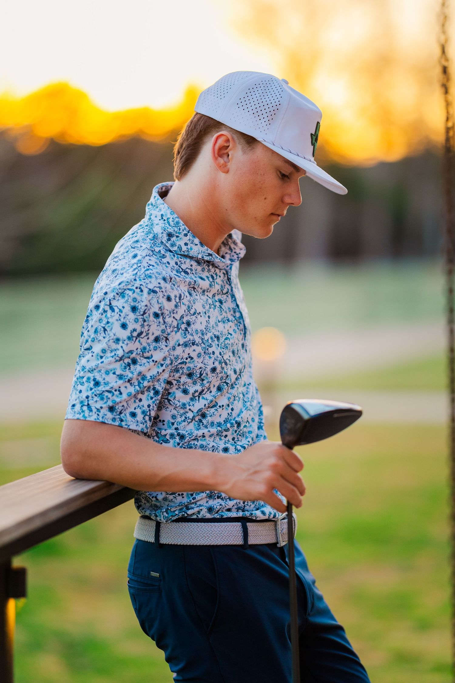 Teen golfer in floral polo shirt and white cap holds golf club leaning on rail at sunset.