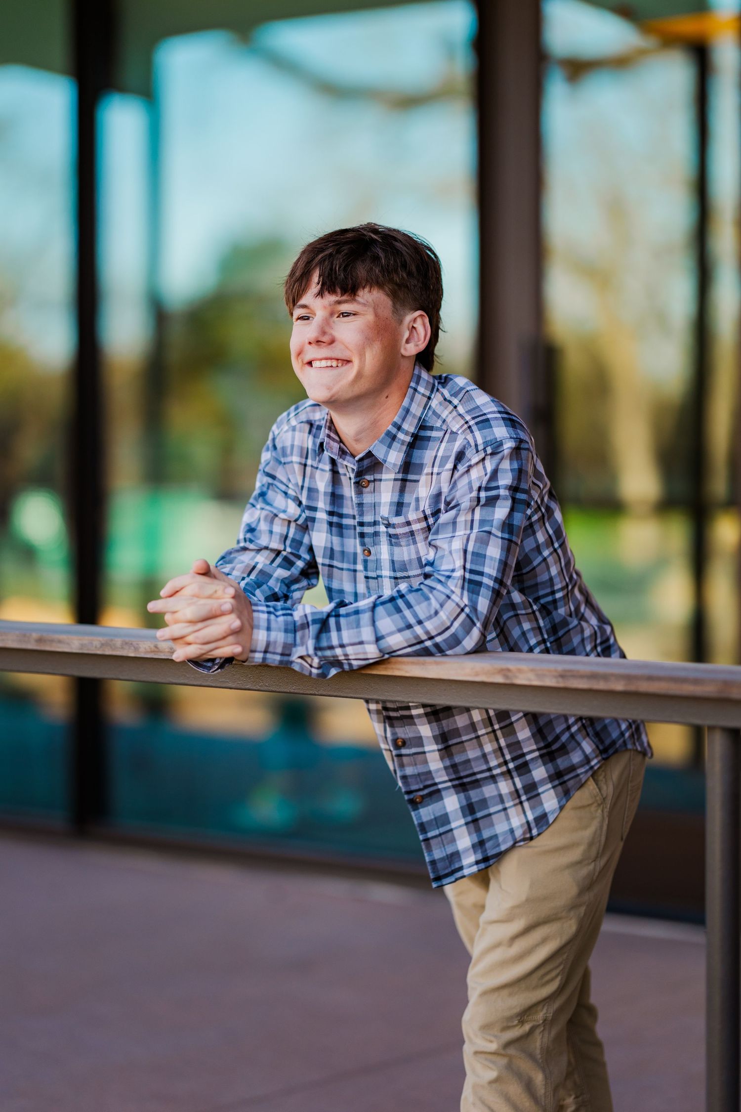 Happy teenage boy in plaid shirt leaning on railing outside glass building smiling.