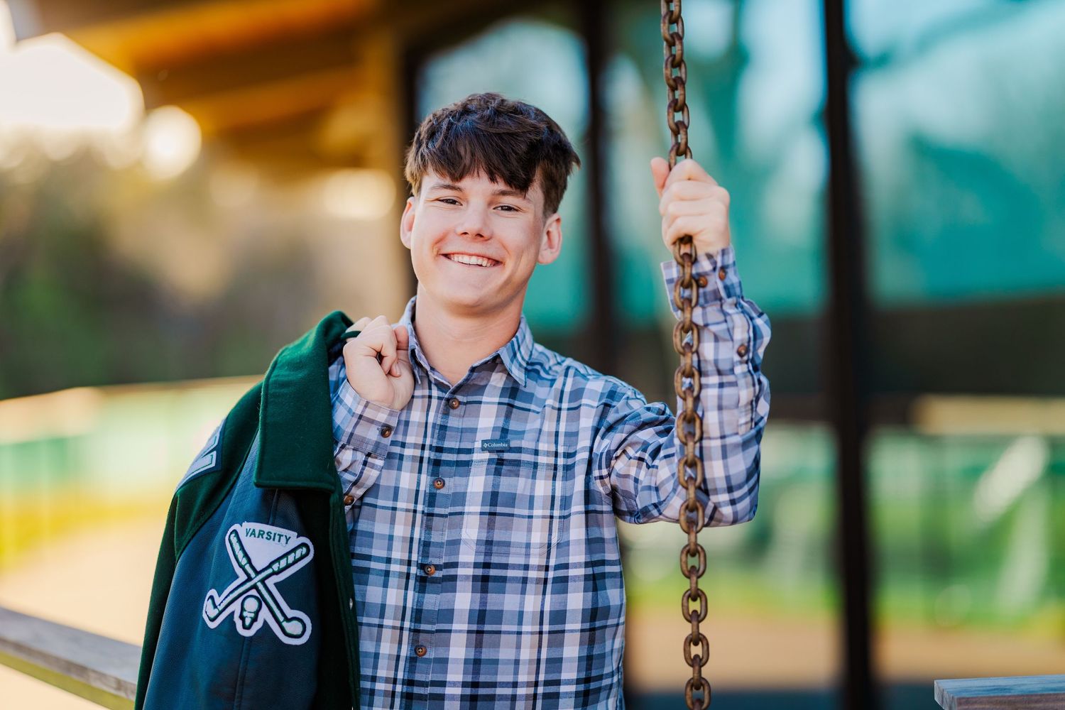 Smiling teen boy in plaid shirt holds chain wearing green varsity jacket in golden hour light.