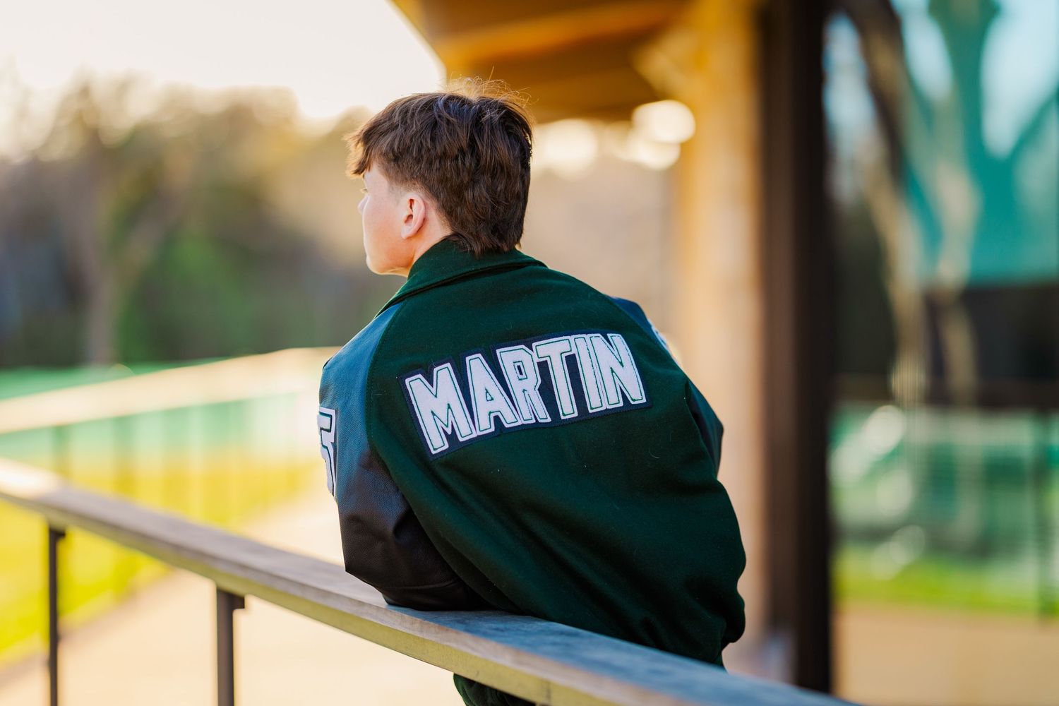 Teen in green Martin varsity jacket sitting at bleachers viewed from behind outdoors.