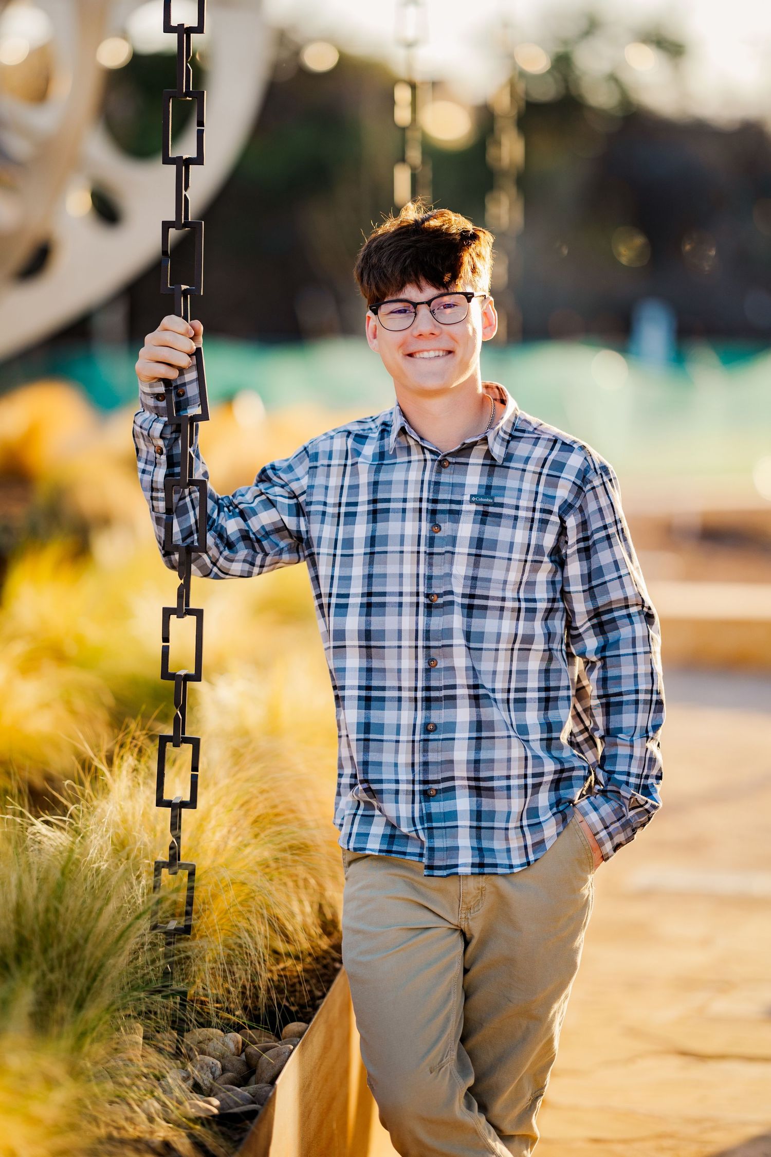 Smiling teen in plaid shirt holding chain sculpture at golden hour outdoor setting.