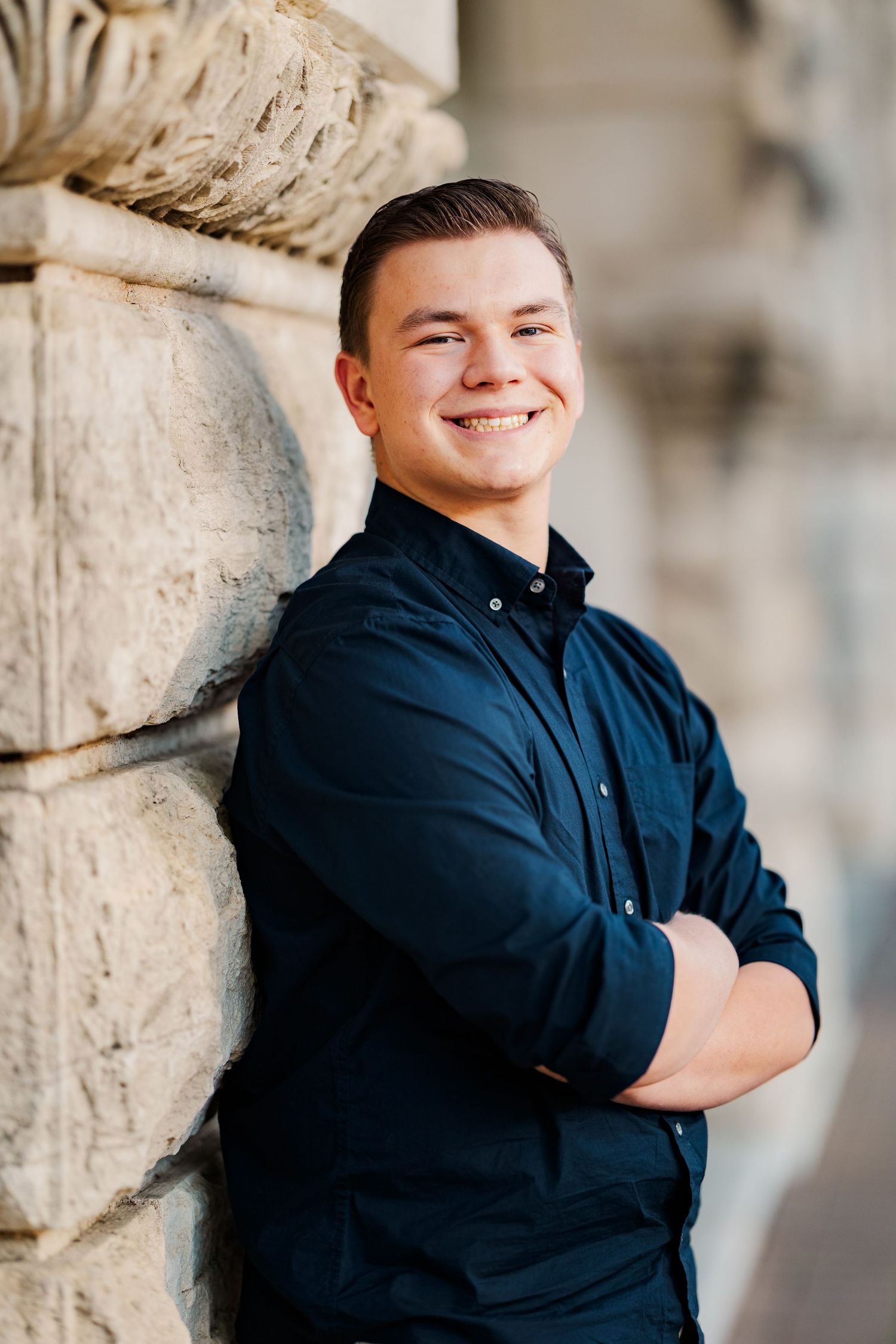 Young man in navy shirt smiling with arms crossed leaning against stone column outdoors.