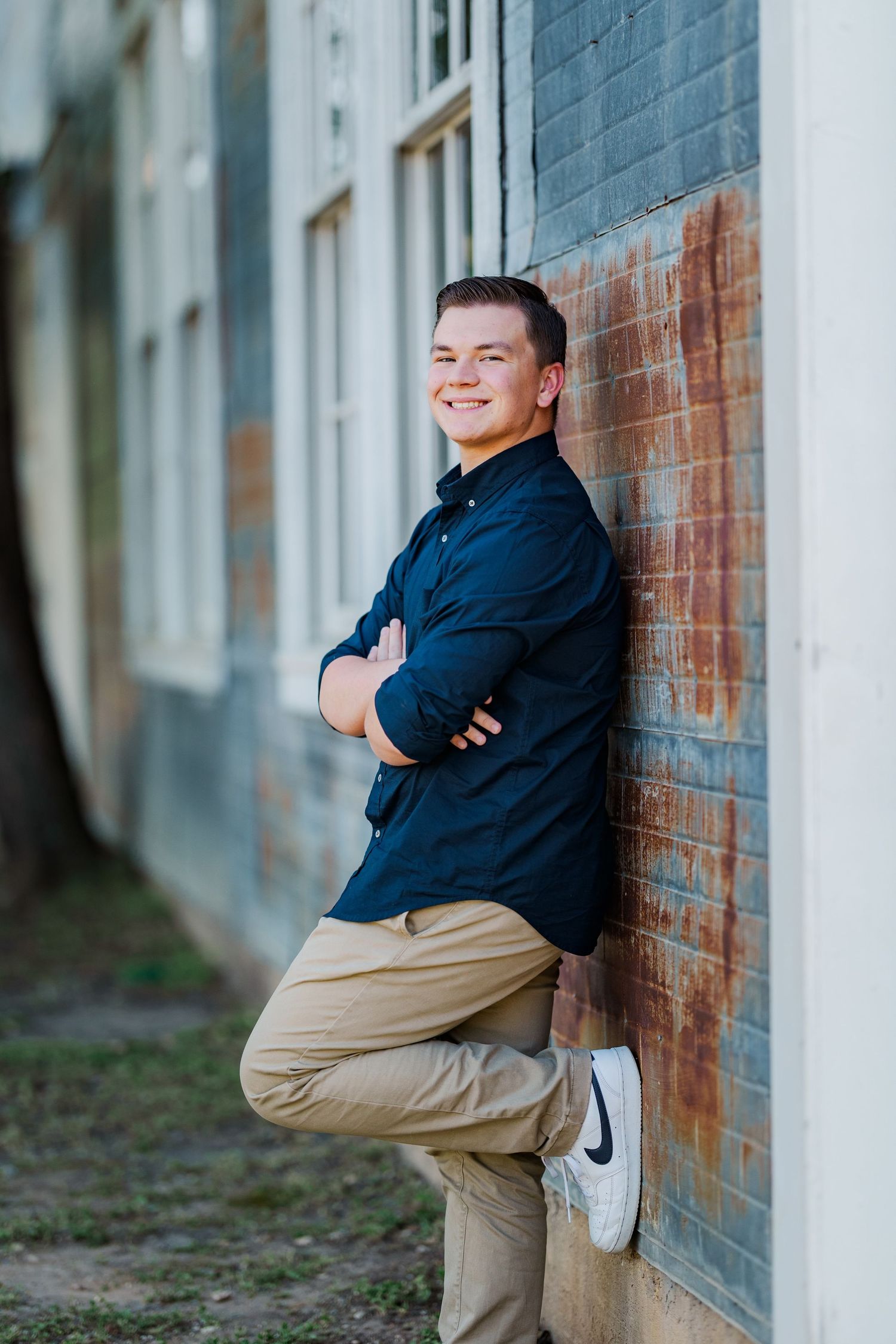 Smiling young man in navy shirt leaning against rustic wall with arms crossed.