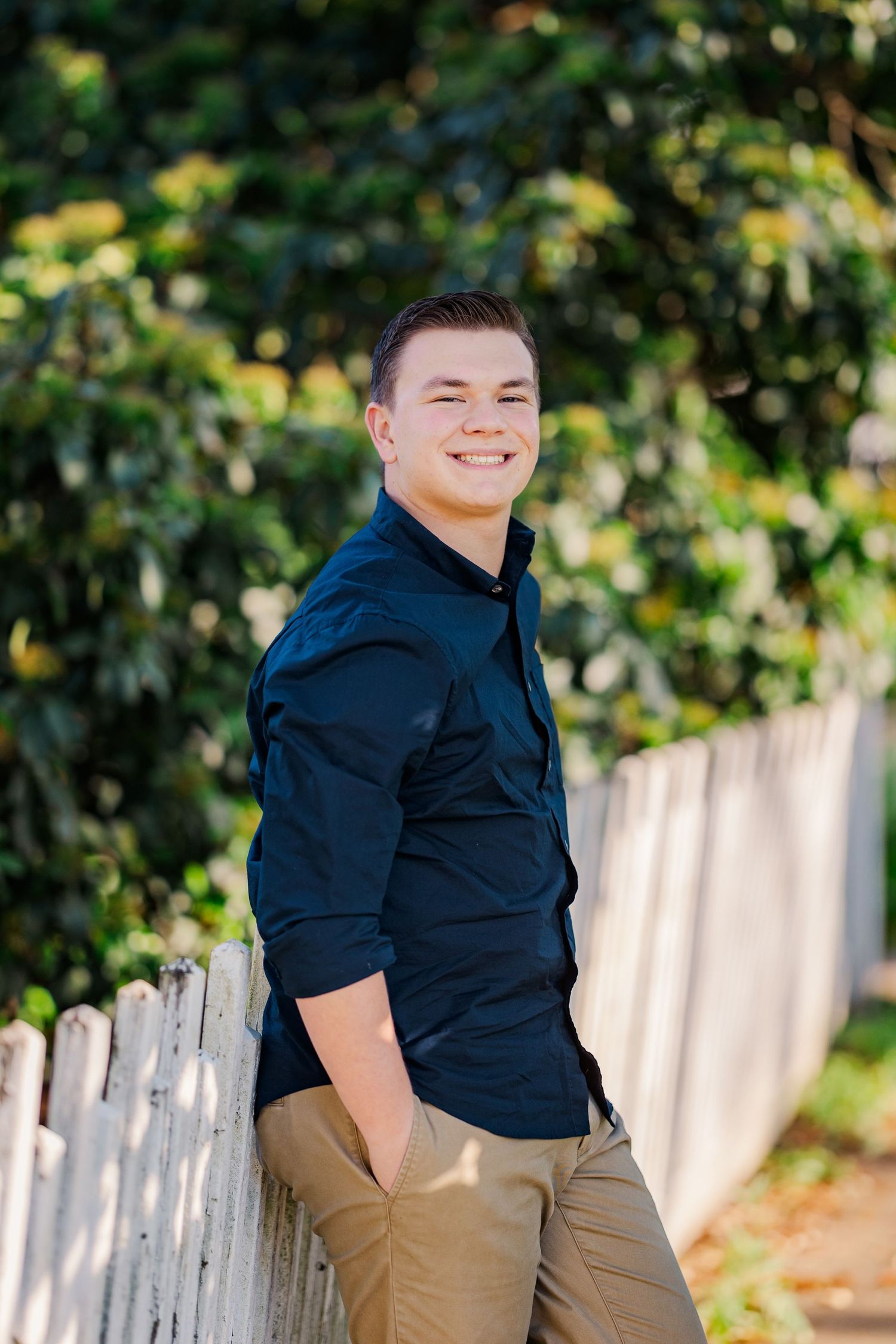Smiling young man in navy blue shirt leaning against white picket fence for outdoor senior photo.