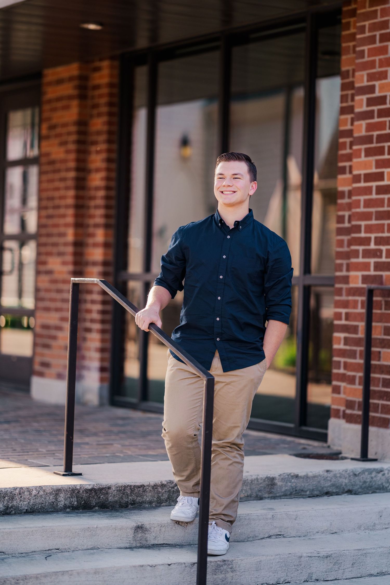 Smiling young man in navy shirt holding railing on brick building steps outdoors.