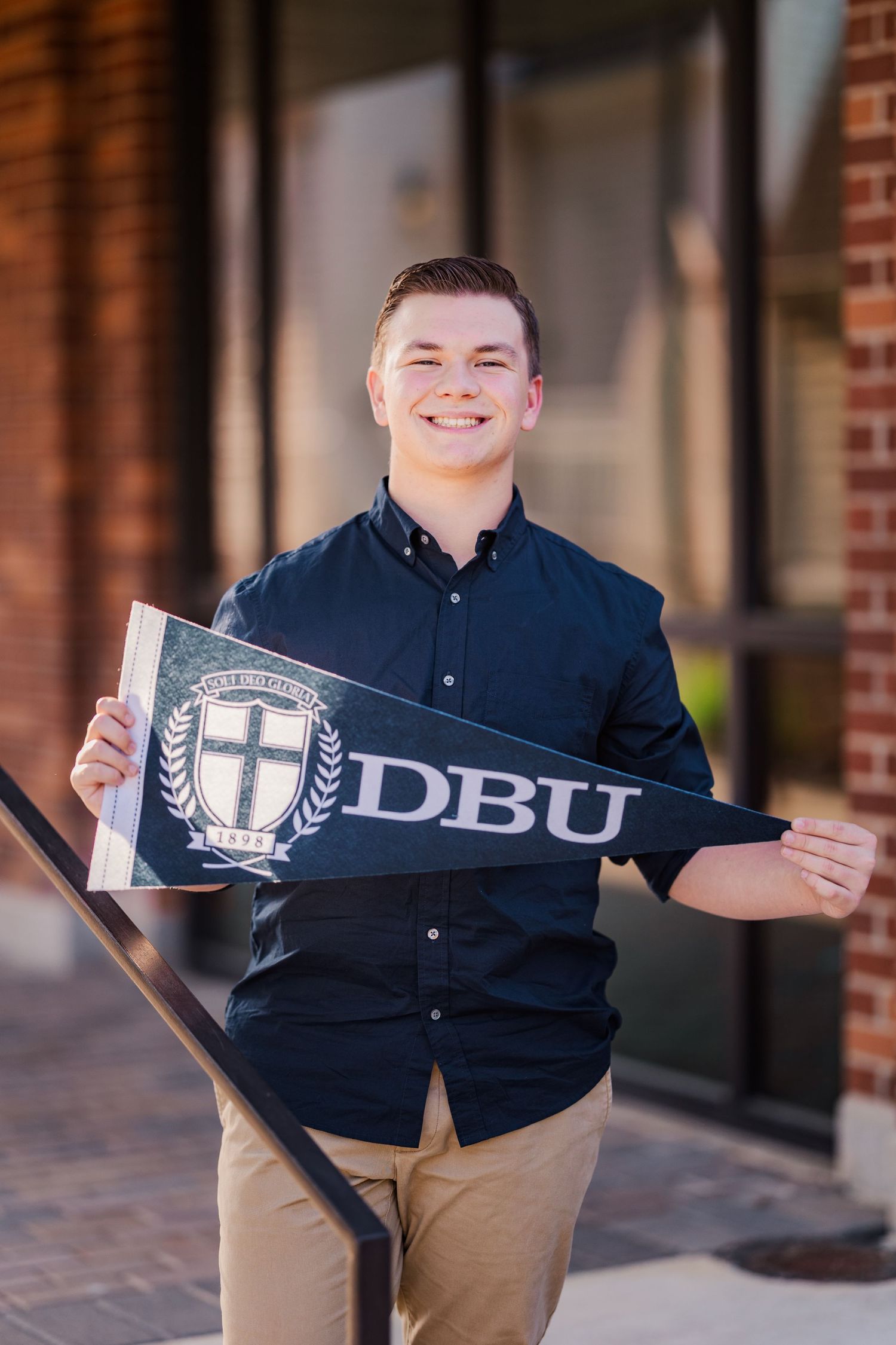 Smiling young man holds a DBU college pennant flag outside a brick building on campus.