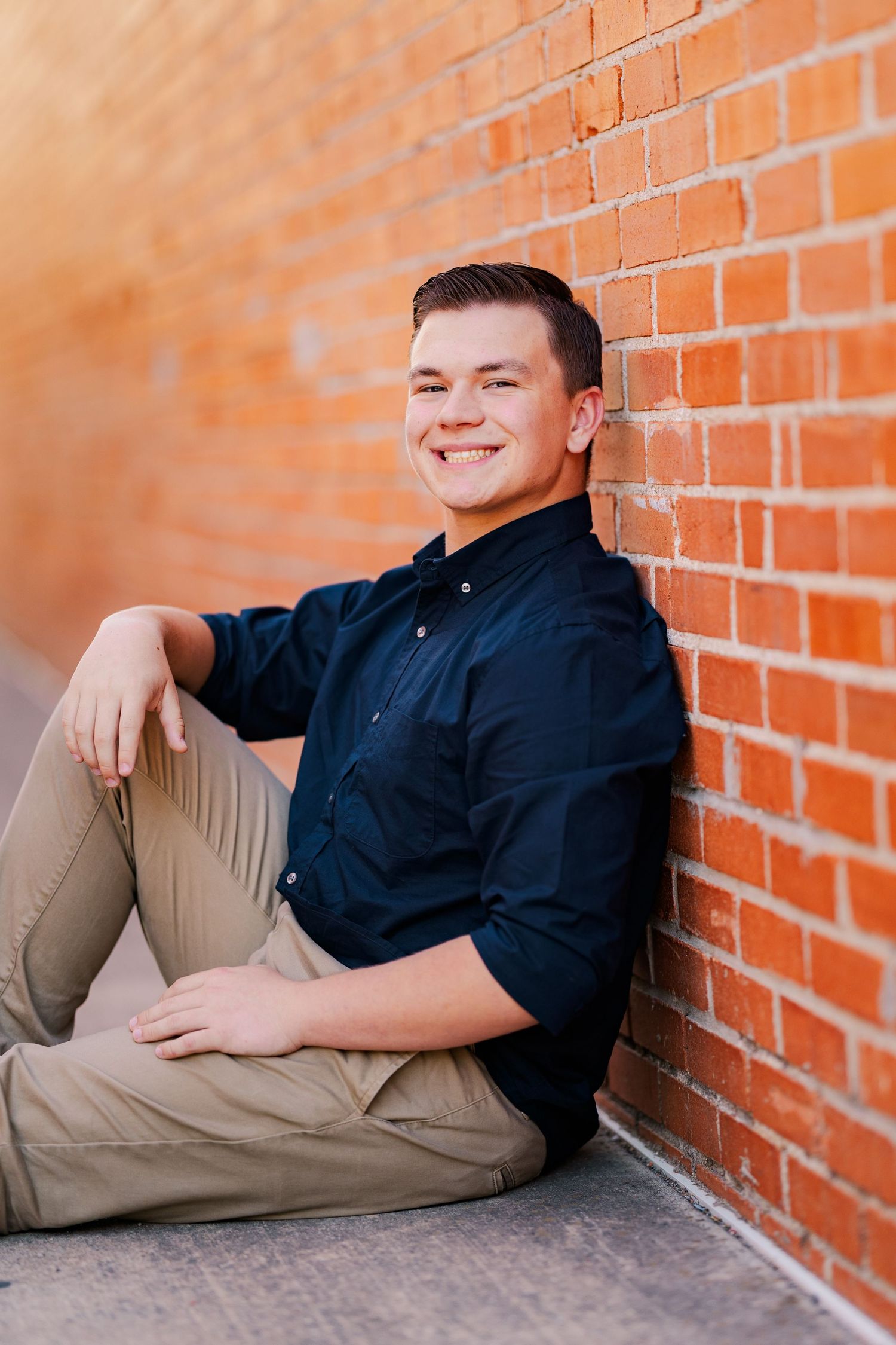 Smiling young man in navy shirt sitting against a red brick wall outdoors.