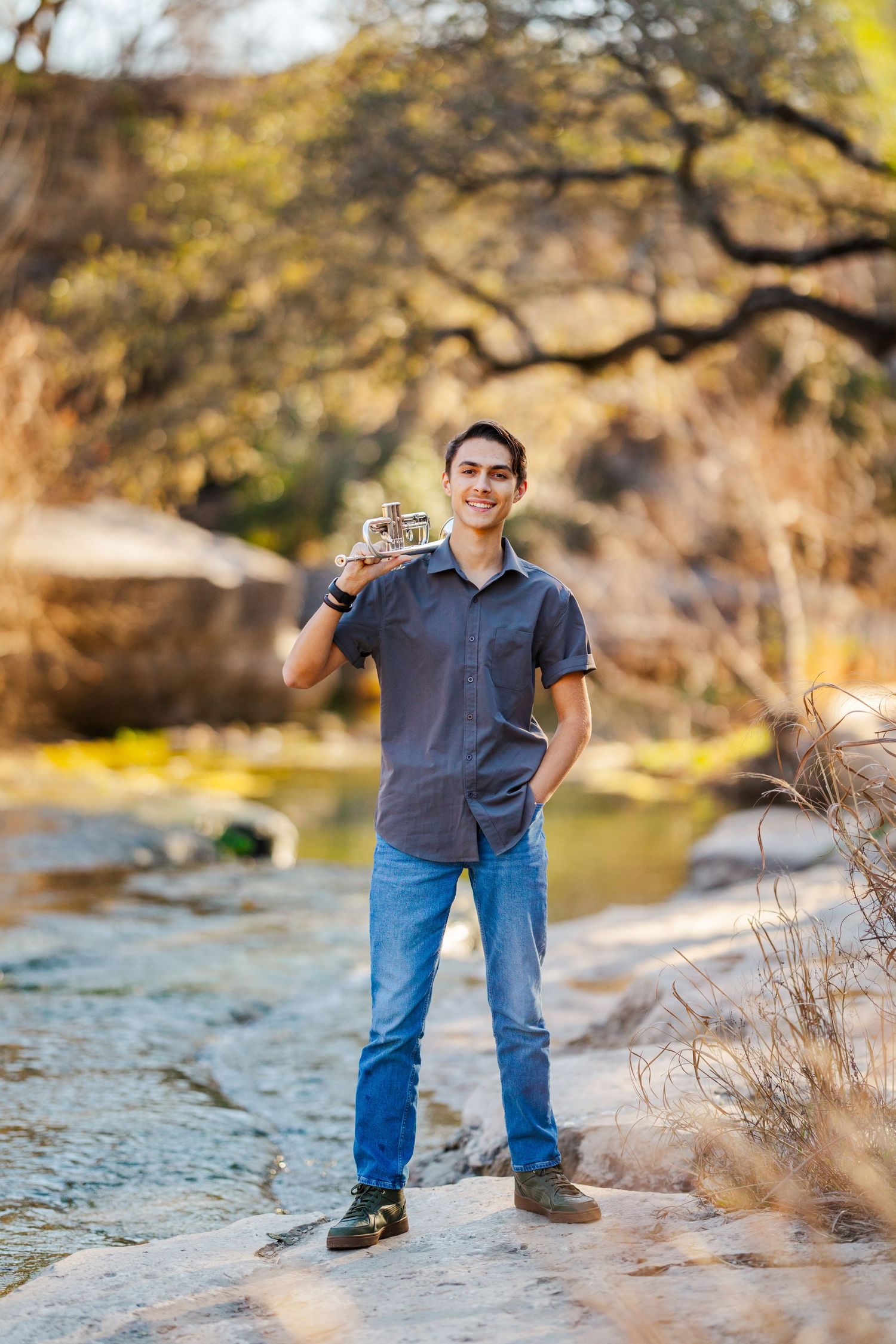 Smiling young man holds a vintage camera while standing on rocks near a creek in a natural outdoor setting.