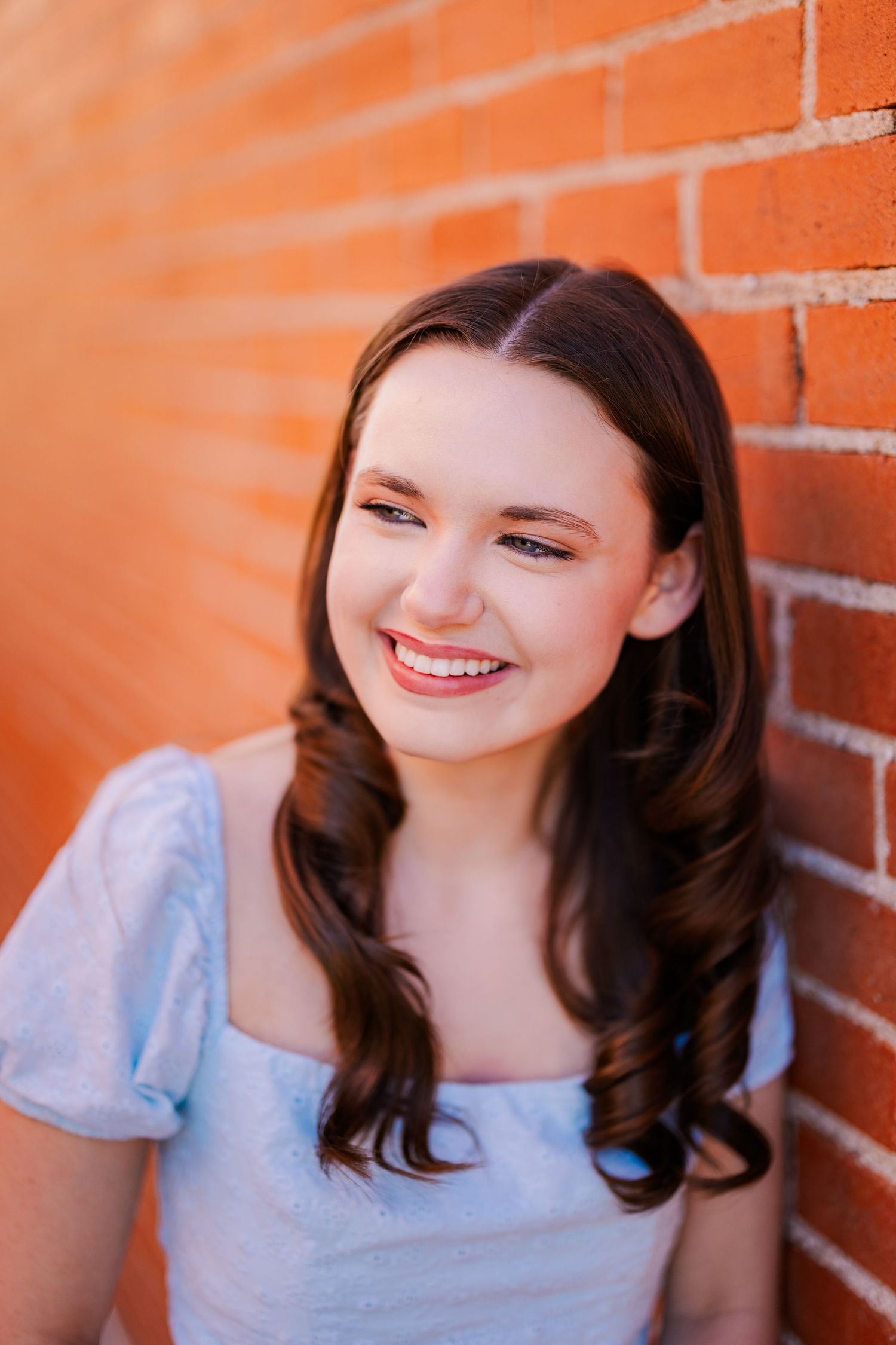 Brunette teenage girl in light blue top laughing against red brick wall for senior portrait.