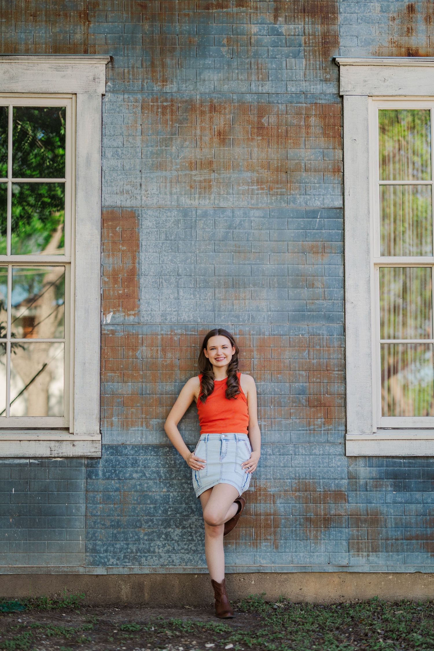 Young woman in orange top and denim skirt leans against rustic weathered blue corrugated metal wall.