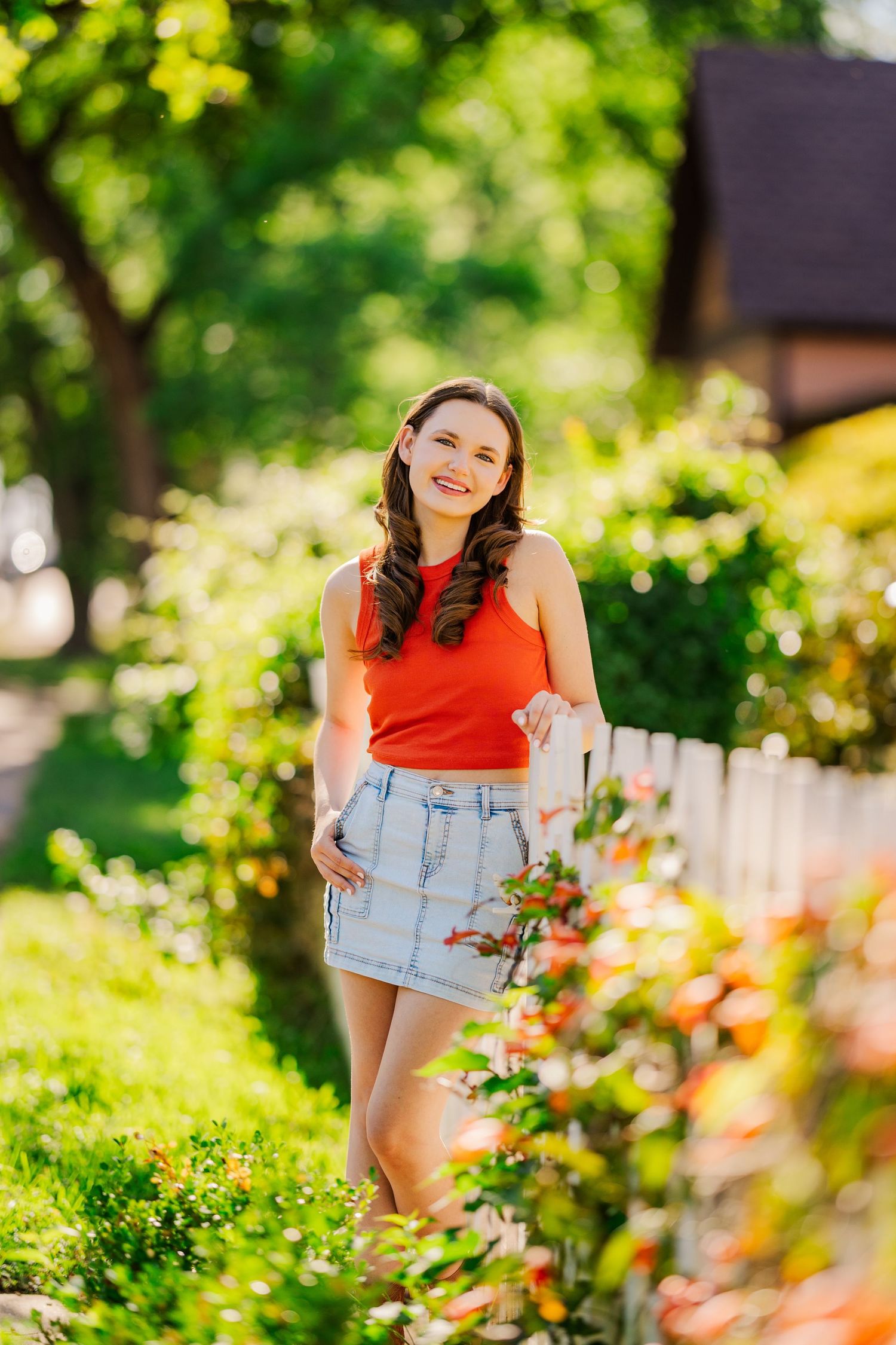 Young woman in red top and denim skirt poses by colorful garden flowers in bright summer sunlight.