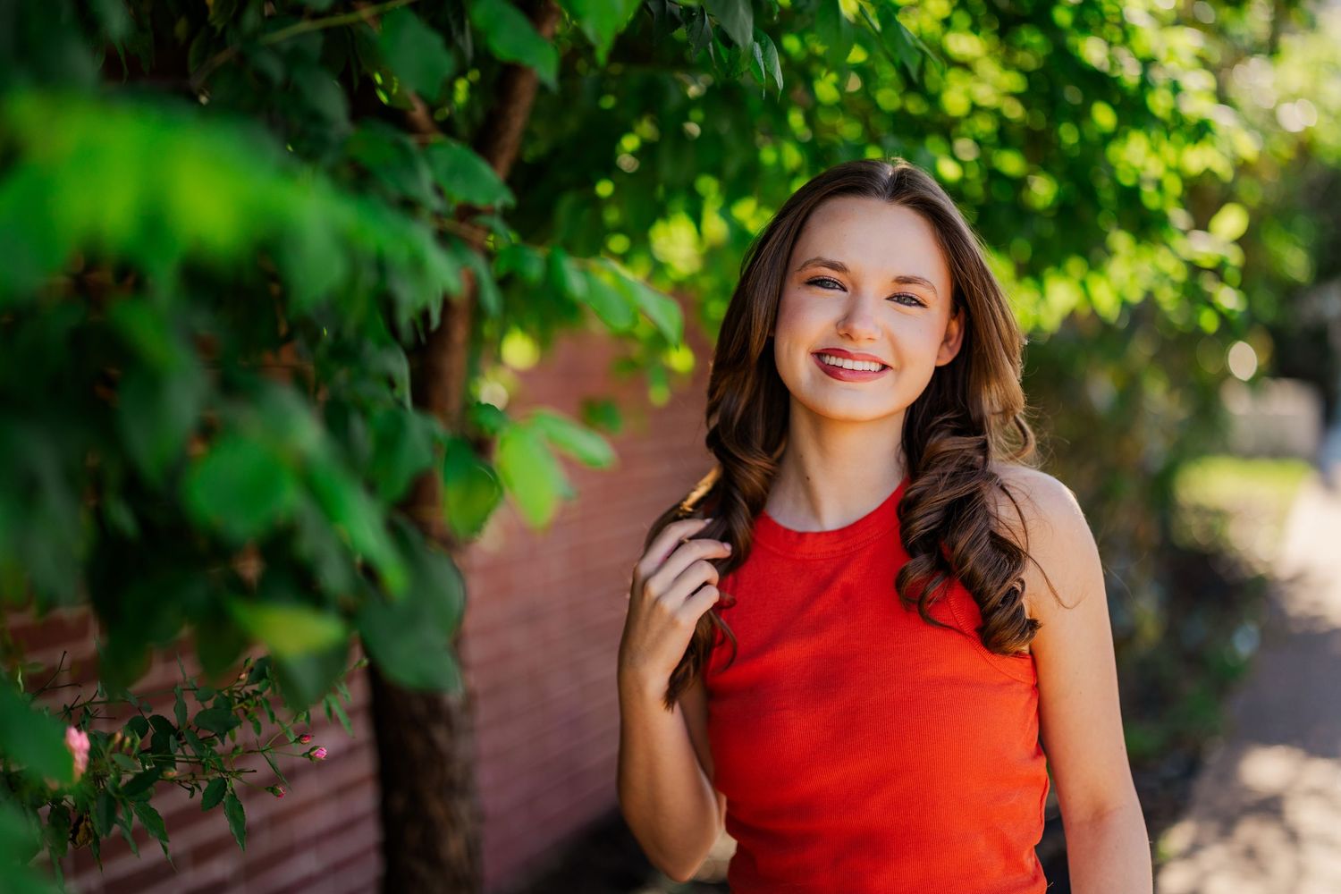Smiling brunette woman in red top posing outdoors near green ivy-covered brick wall.