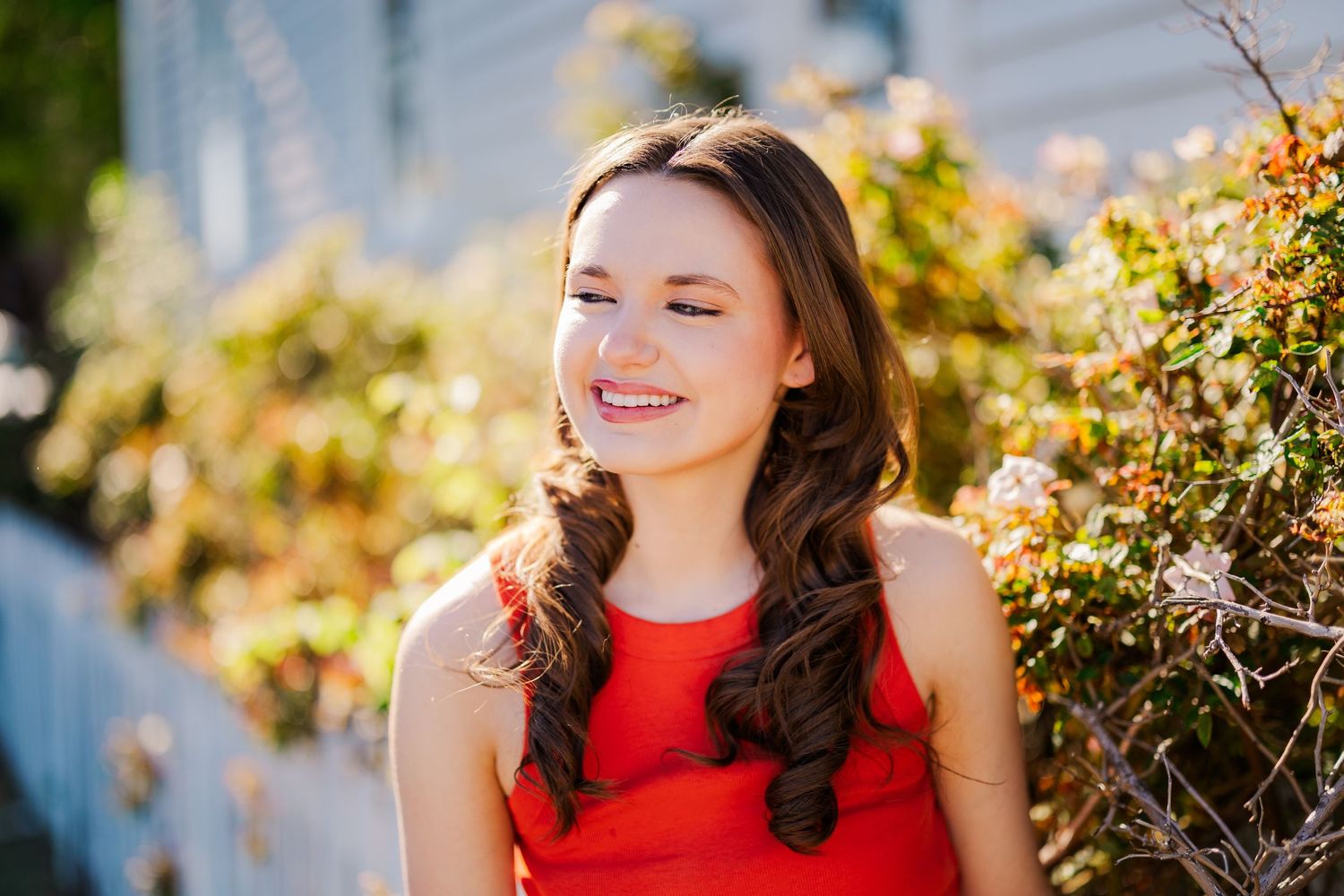 Smiling teenage girl with long brown hair in red dress outdoors in sunny garden.