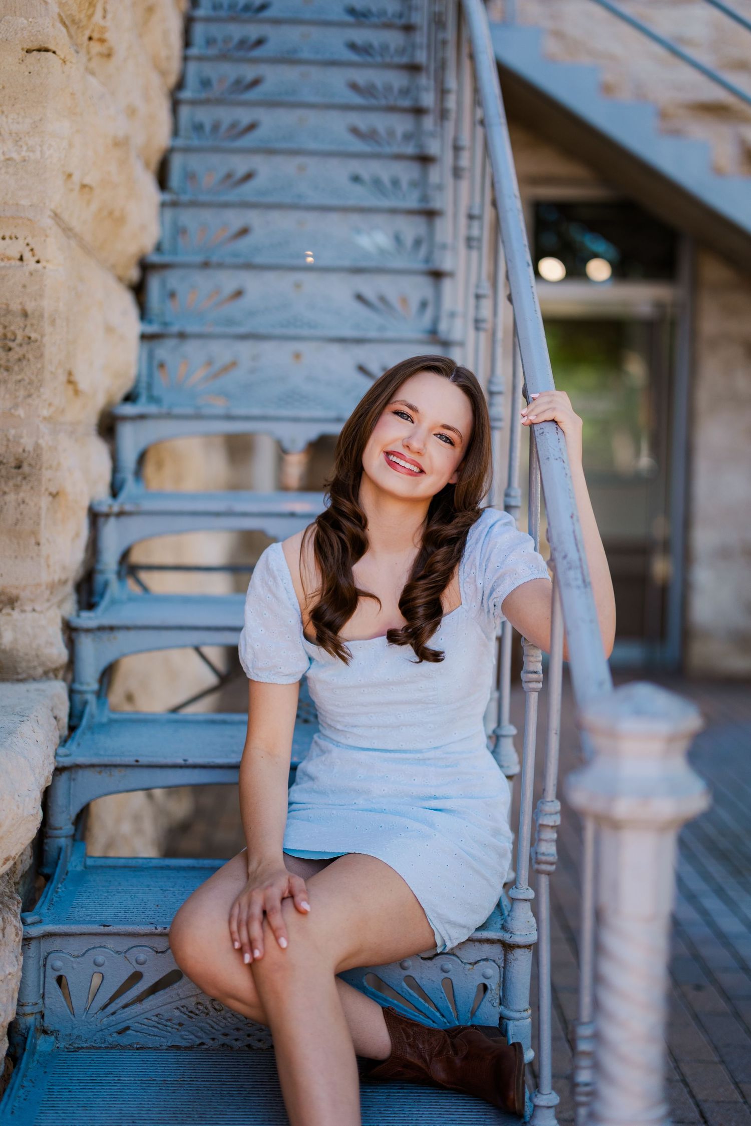 Smiling young woman in white dress sitting on vintage blue iron staircase outdoors.