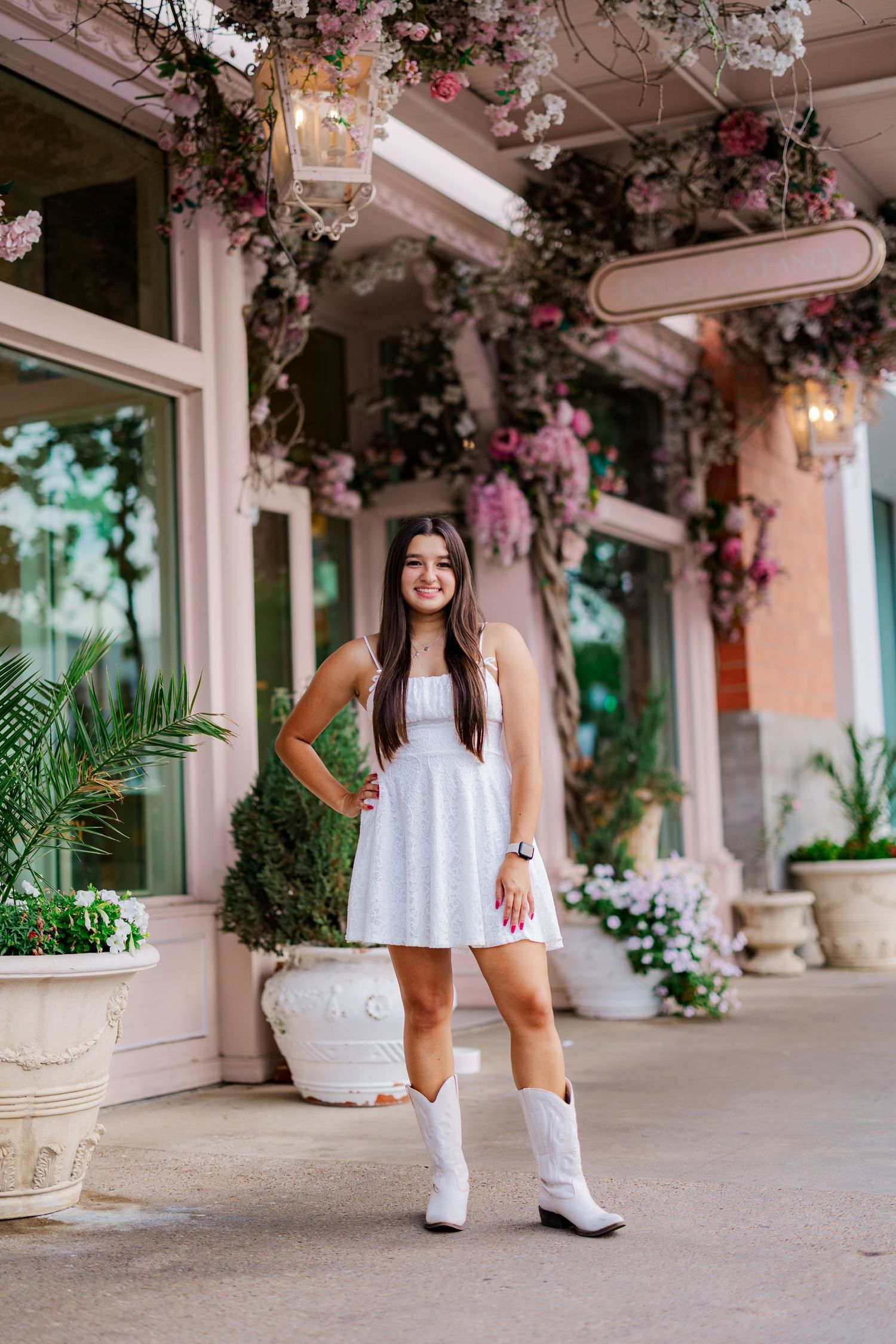 Young woman in white dress and cowboy boots posing outside floral-decorated pink boutique.