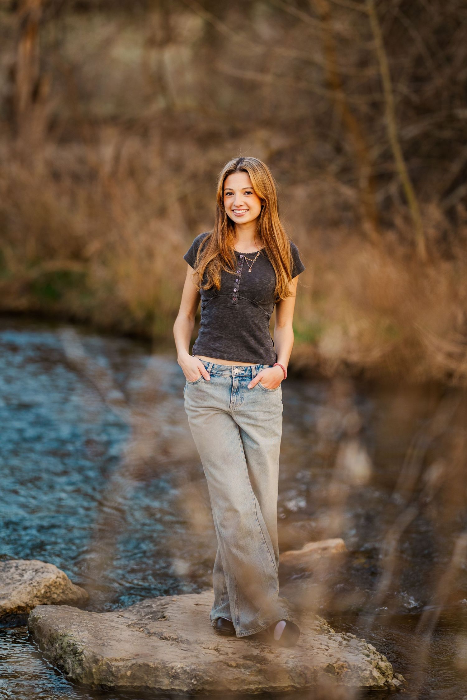 Smiling teen girl in gray tee and wide-leg jeans stands on rocks beside a peaceful woodland creek.