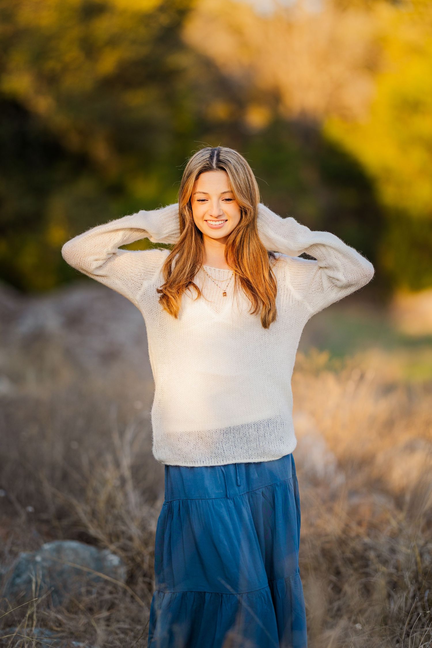 Smiling teen girl in cream sweater and blue skirt poses in a sunlit autumn outdoor setting.