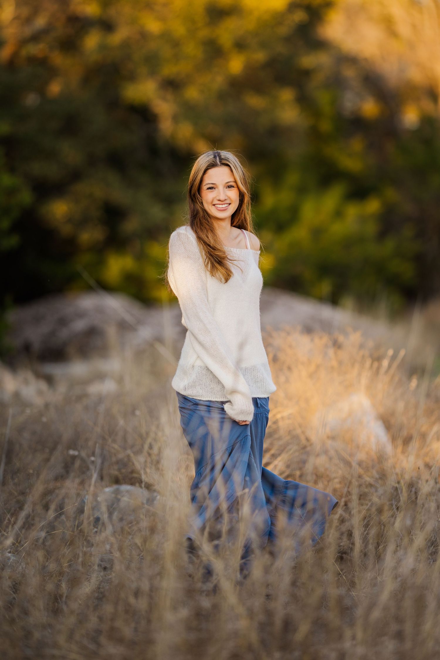 Smiling young woman in white sweater and jeans stands in golden dry grass at sunset.