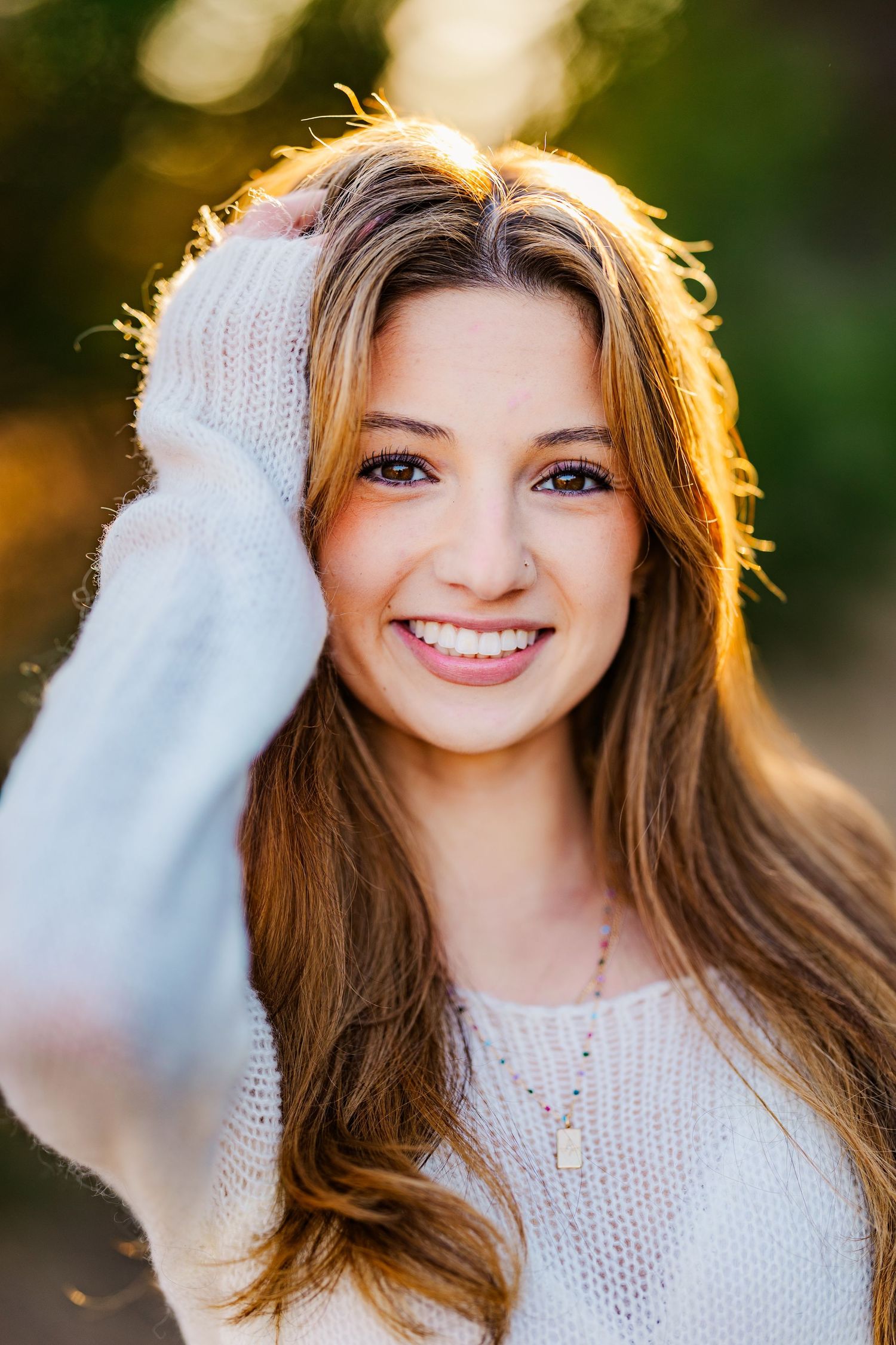 Beautiful smiling teenage girl with long hair in white sweater poses outdoors in golden sunlight.