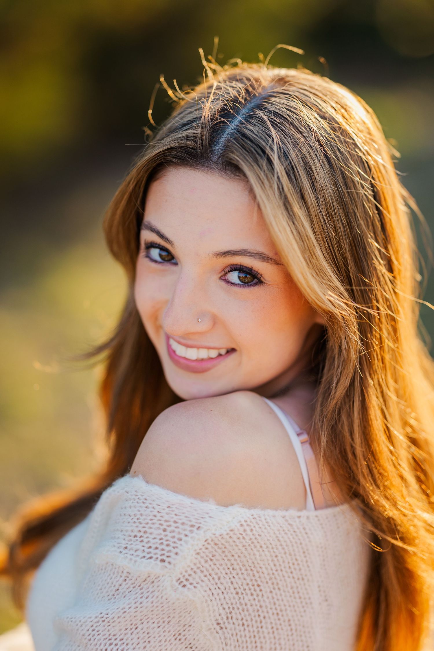 Young woman with long brown hair smiles over shoulder in warm golden sunlight wearing off-shoulder cream top.