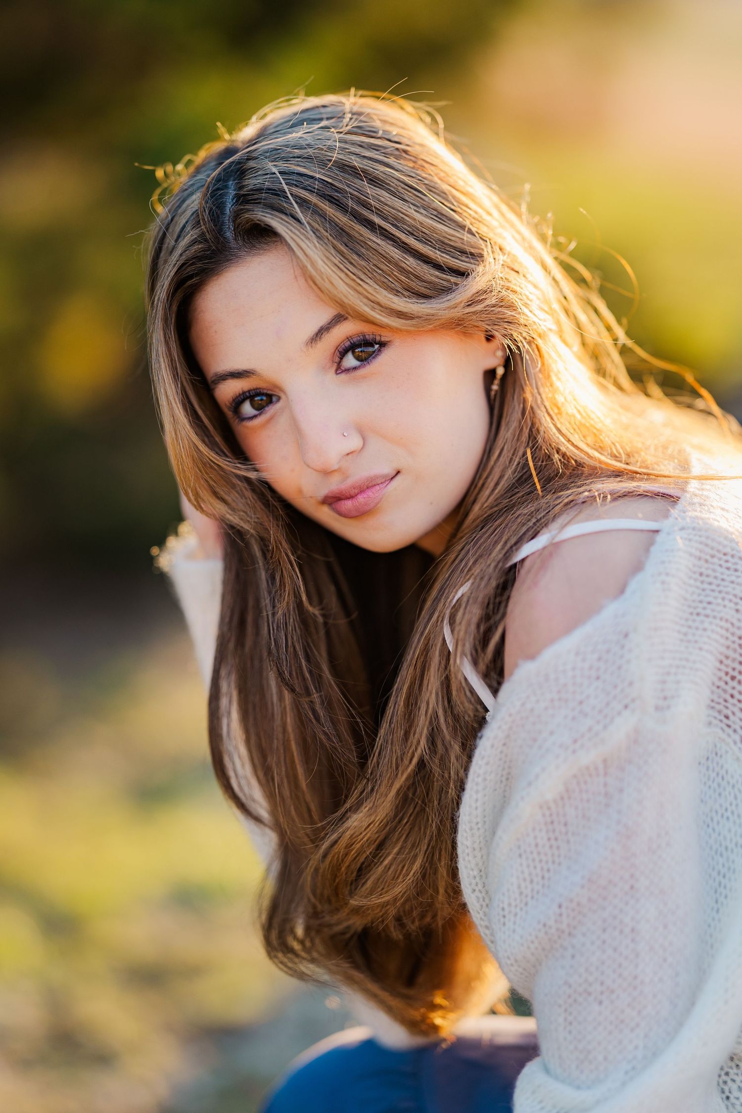Beautiful young woman with long brown hair posing outdoors in warm golden sunlight.