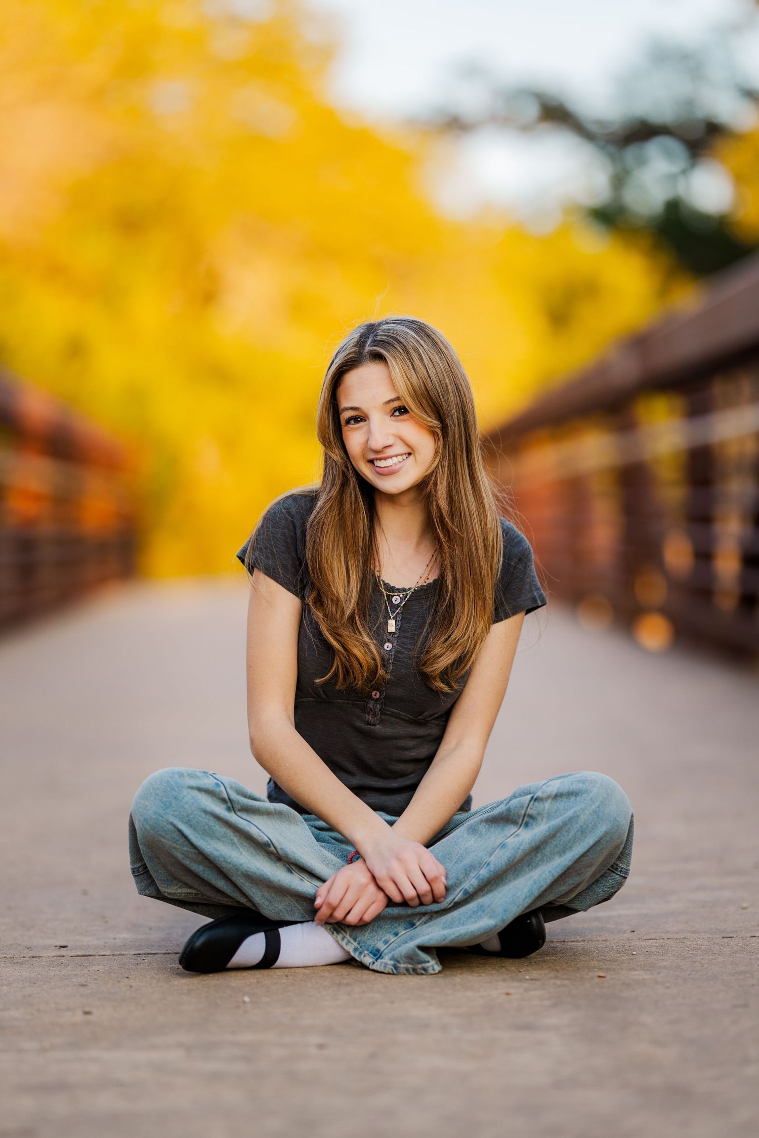 Smiling teenage girl in casual outfit sits cross-legged on a bridge with yellow autumn foliage.