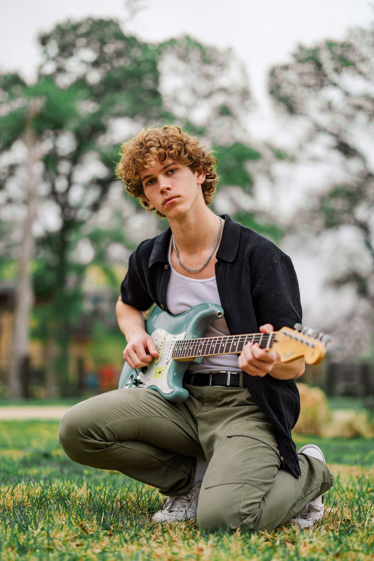 Curly-haired teen boy kneeling on grass playing an electric guitar in an outdoor park.