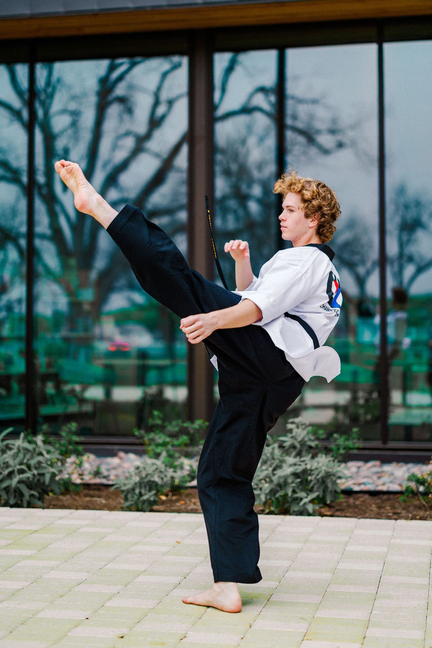 Curly-haired teen in martial arts uniform performs a high kick outdoors in front of a glass building.