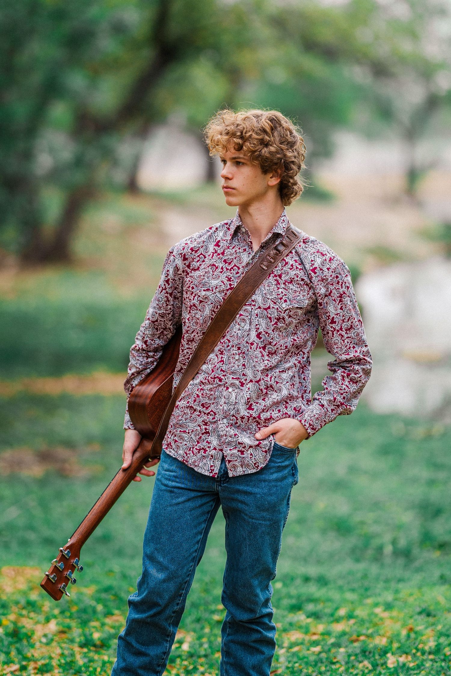Curly-haired teenage boy holding a guitar outdoors in a green park setting wearing a floral shirt.