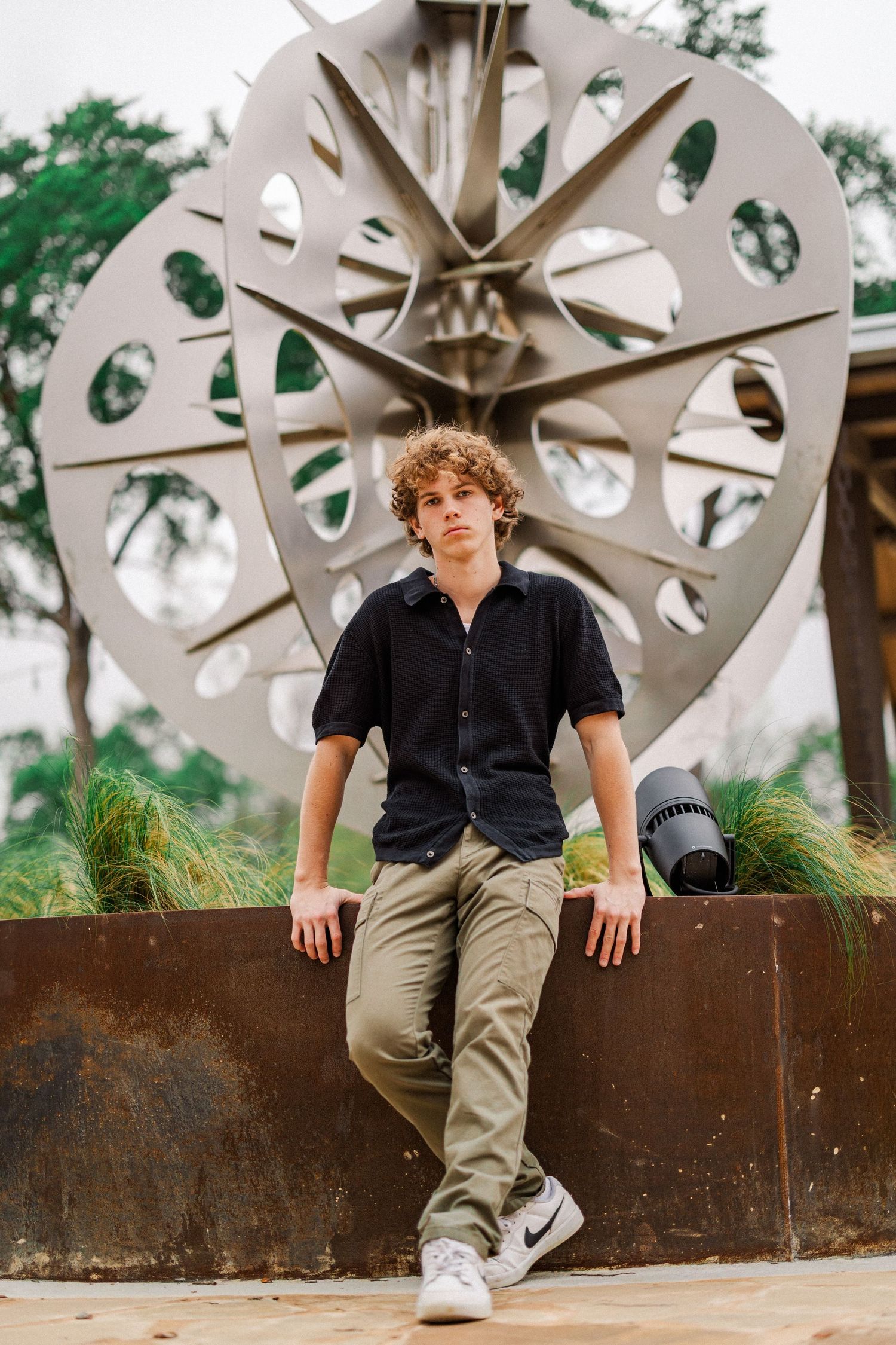 Curly-haired teen boy in black shirt leaning against a wall near a large metal sculpture outdoors.