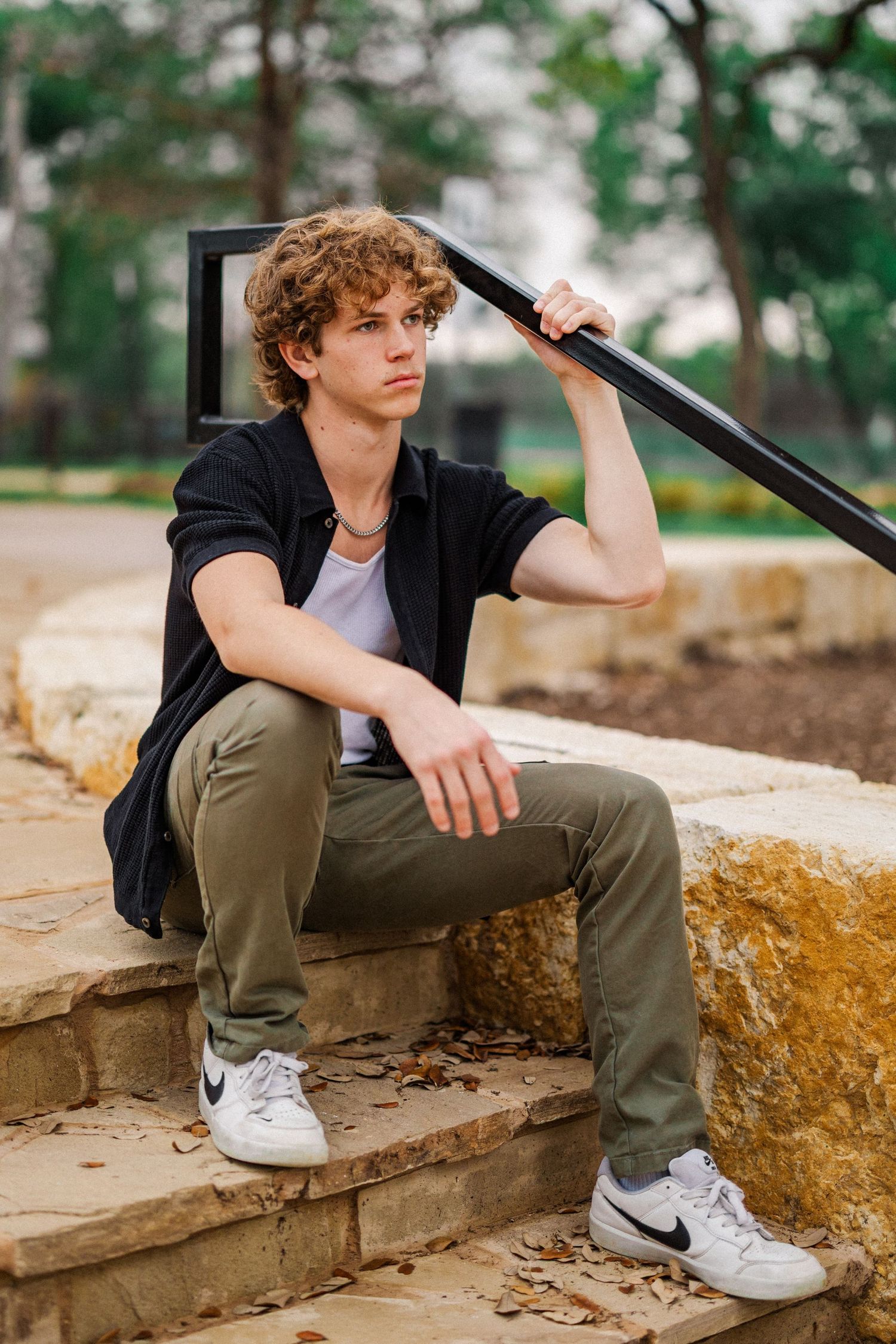 Curly-haired young man in black shirt sits on stone steps holding metal railing in park setting.