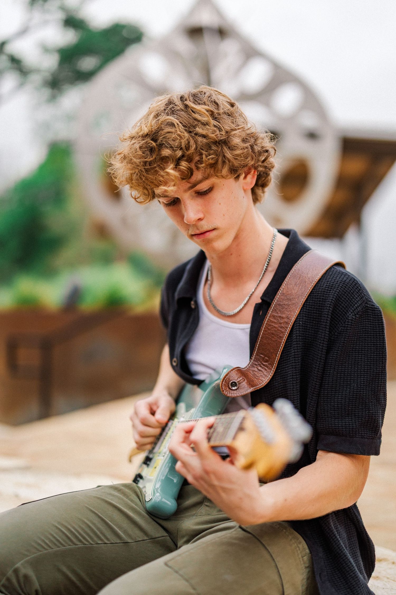 Curly-haired teen boy playing electric guitar while seated outdoors in a garden setting.